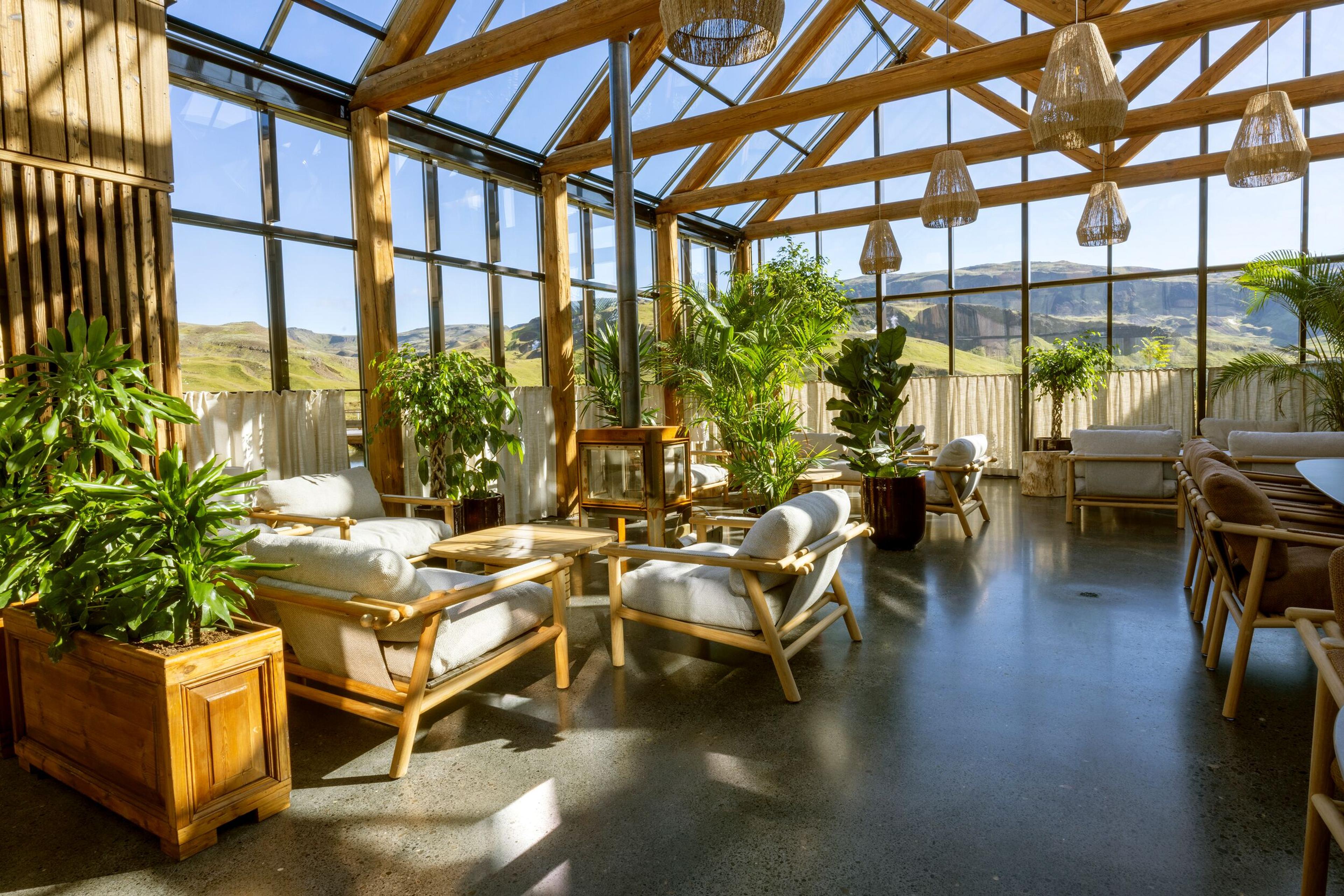 Bright sunroom with a wooden frame, glass roof, many plants, comfortable lounge furniture, and large windows revealing a mountainous landscape.