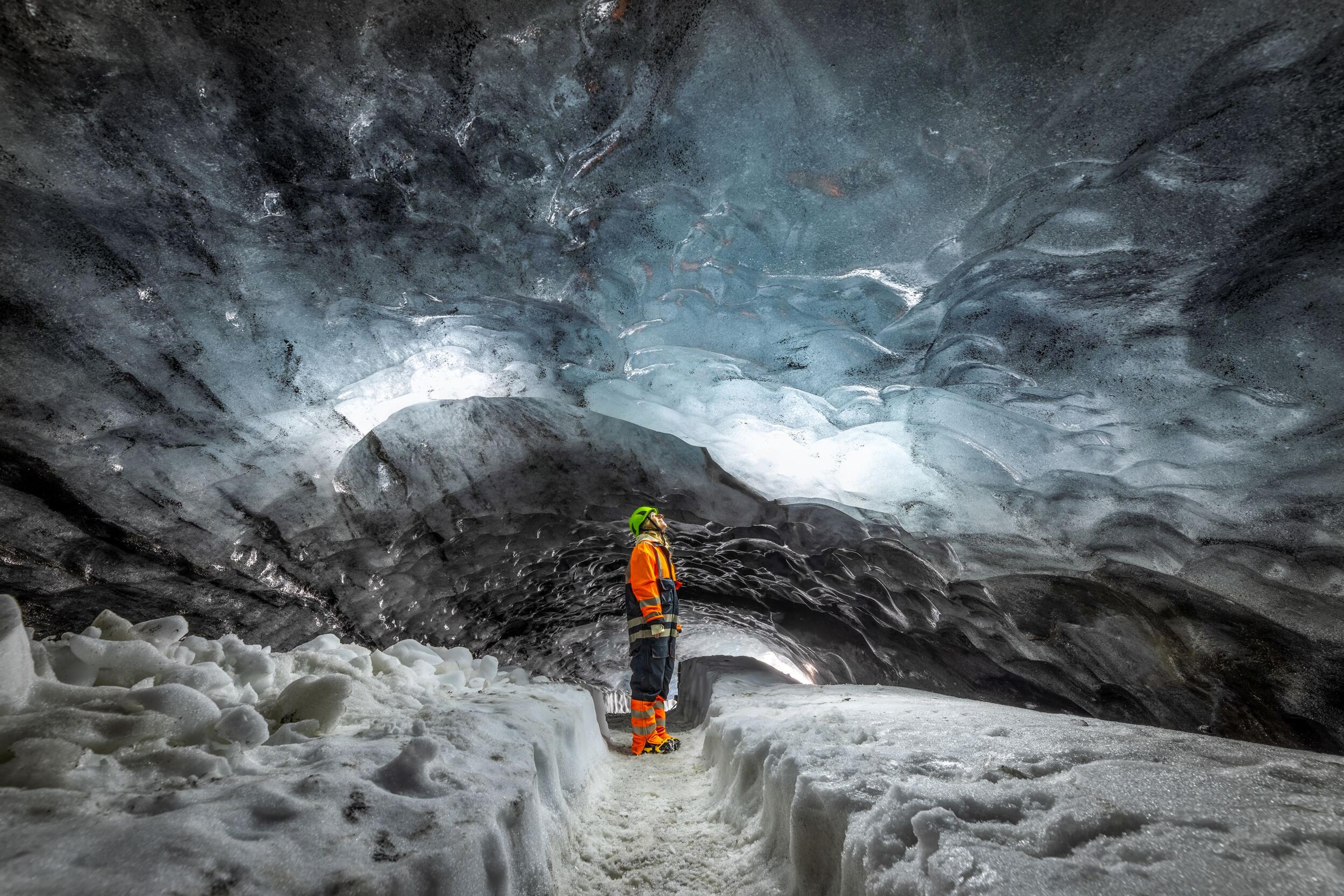 Person in orange and black suit standing in Askur ice cave, surrounded by ice and snow with a glowing ceiling.