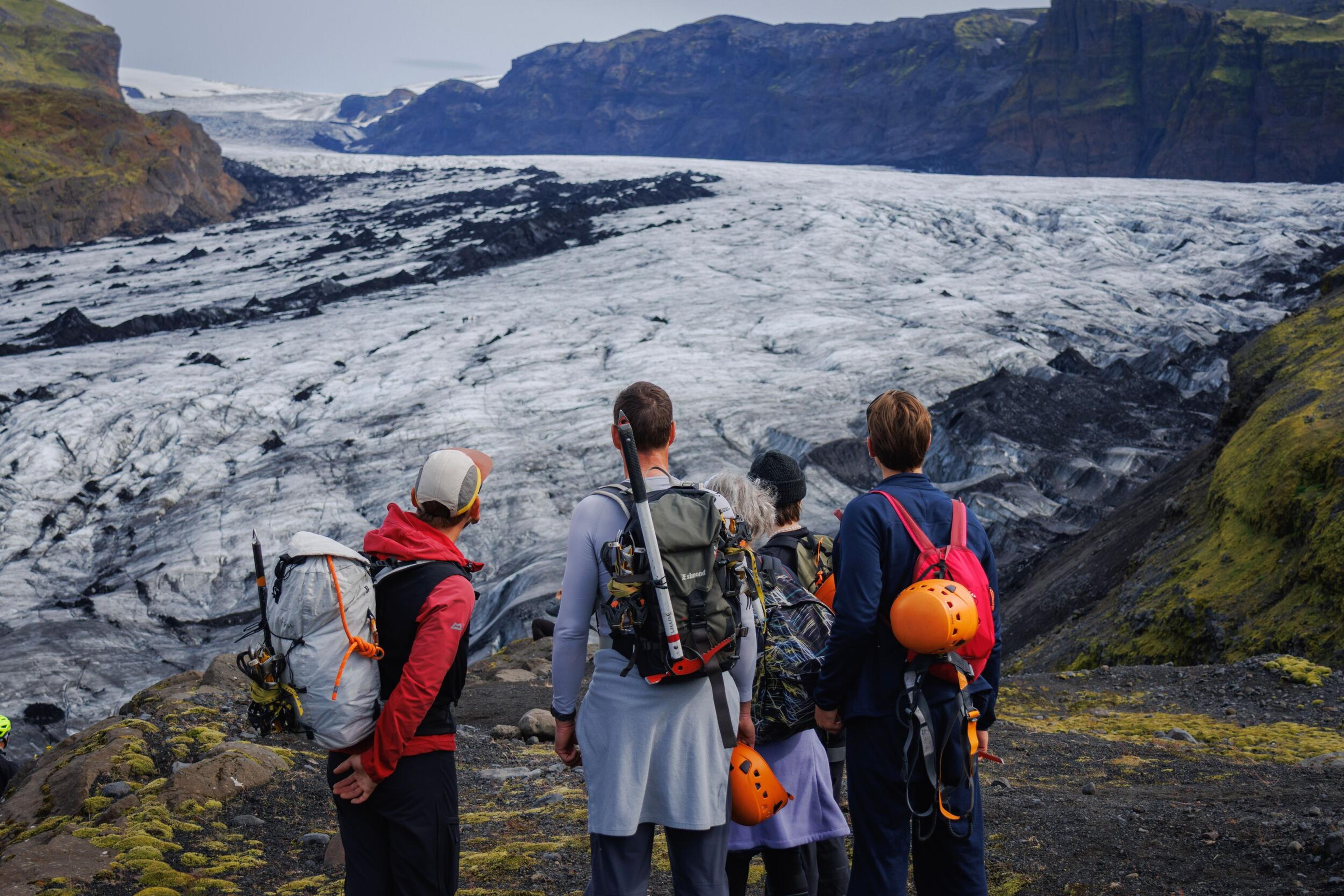 A group of hikers with backpacks looks out over a vast, ash-covered glacier and rocky mountains.