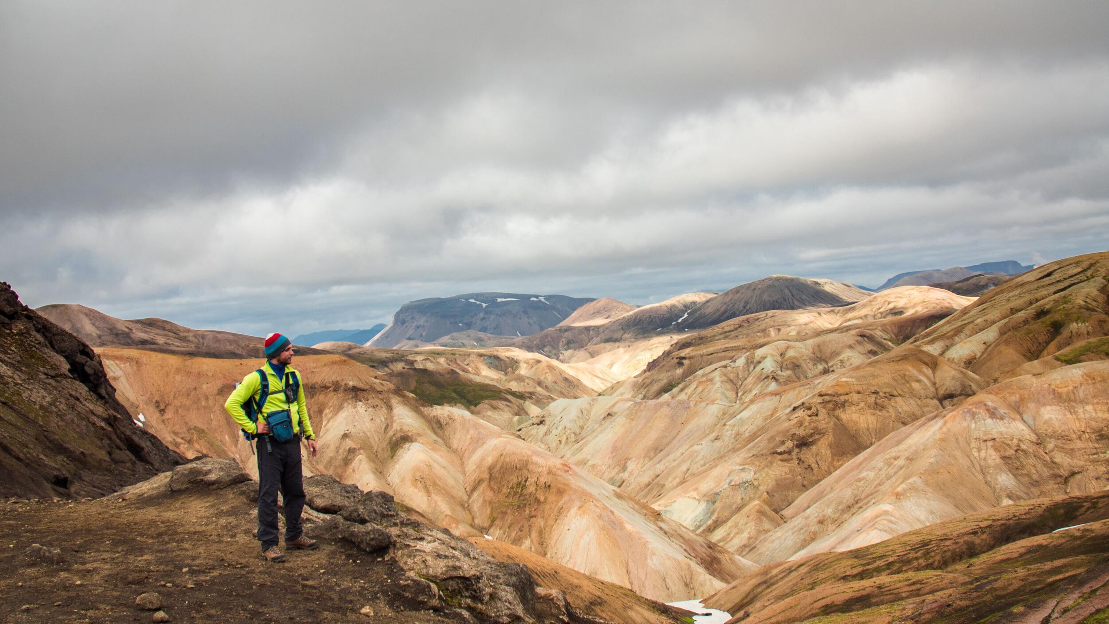 A hiker stands on a ridge overlooking a vast landscape of multicolored mountains under a cloudy sky.