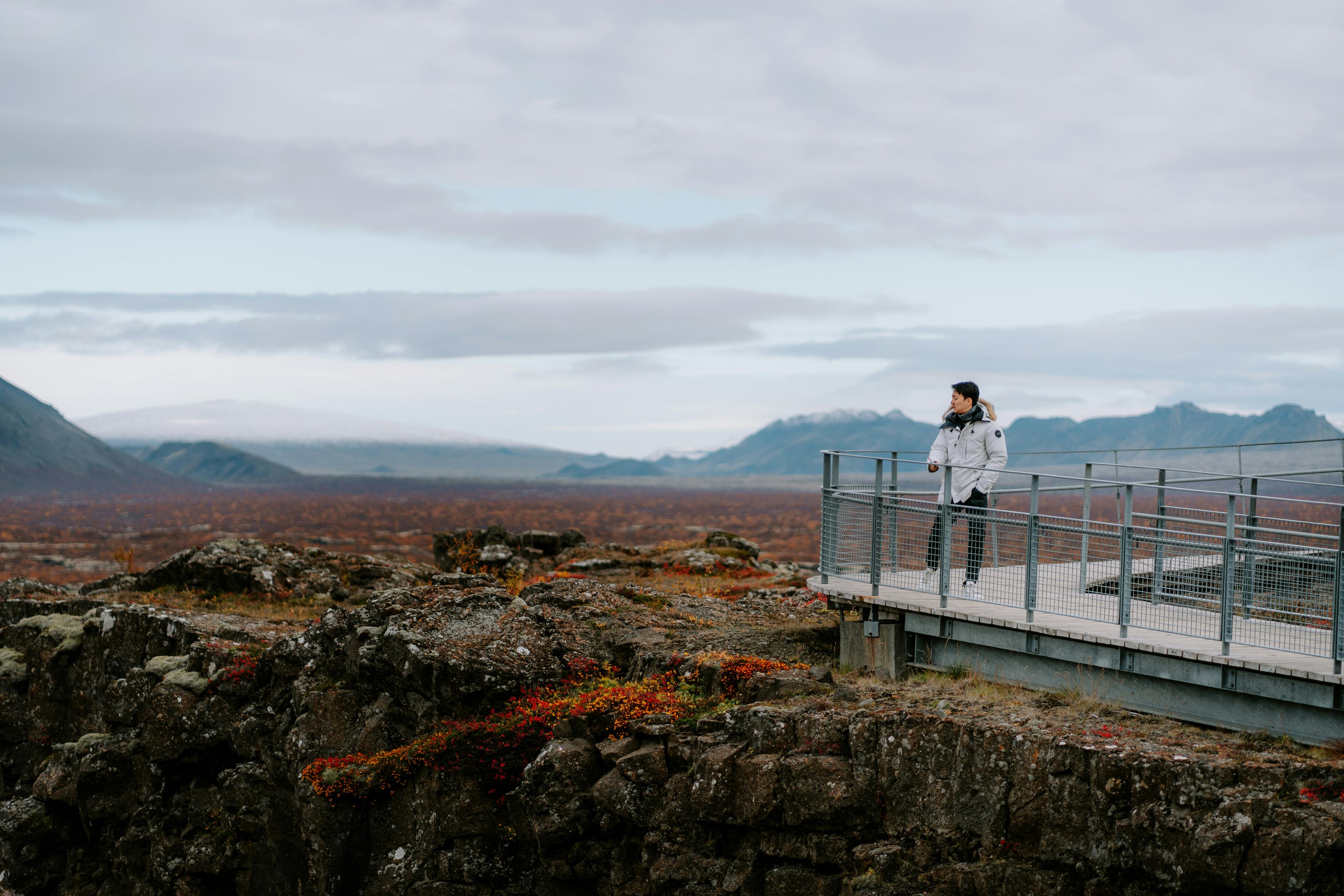 erson on a viewing platform at Thingvellir National Park, Iceland, gazing over a rocky landscape with distant mountains and cloudy skies.