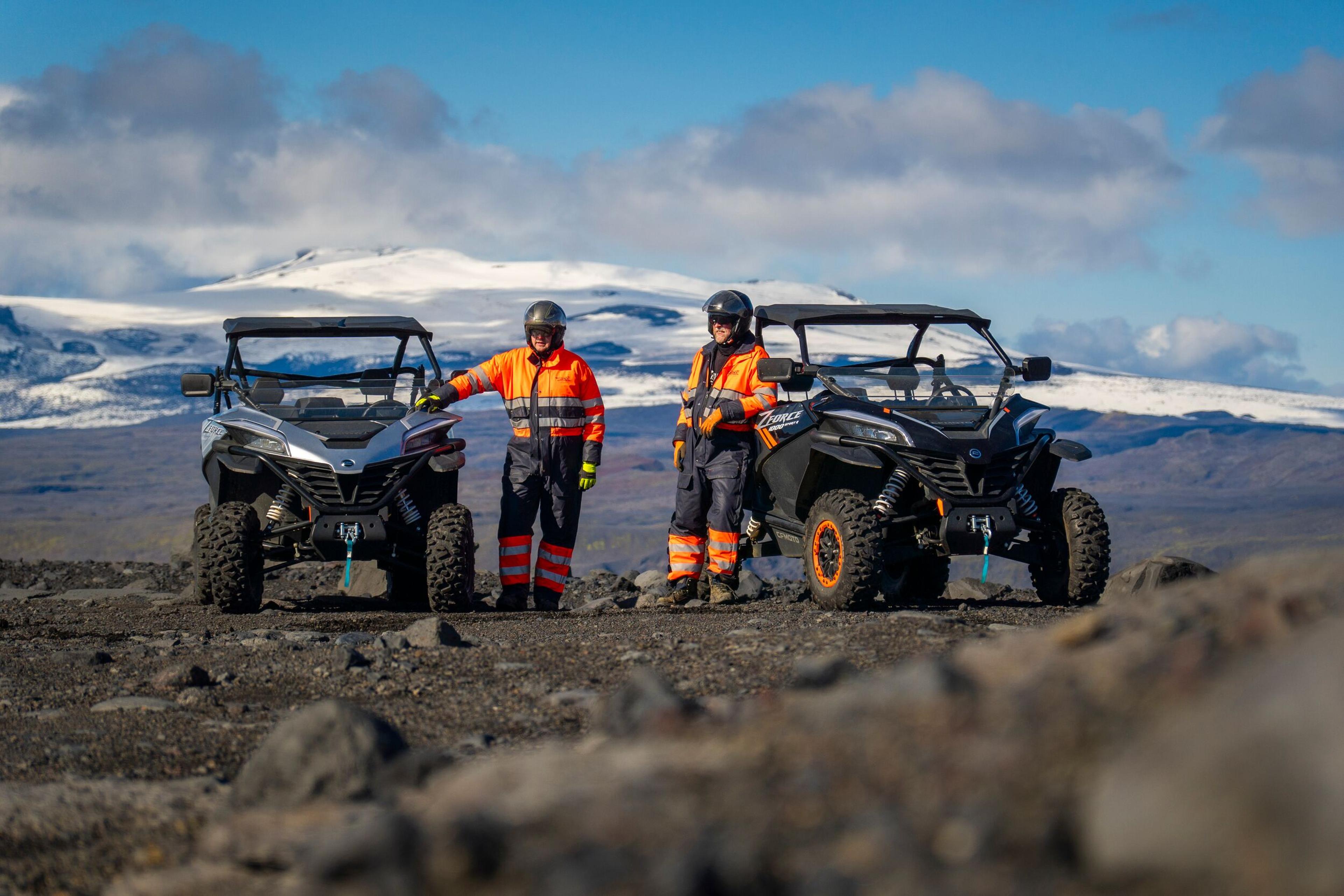 An off-road utility task vehicle (UTV) with two occupants wearing safety gear, including helmets and high-visibility vests, is parked on a rugged terrain with a glacier-topped mountain in the background