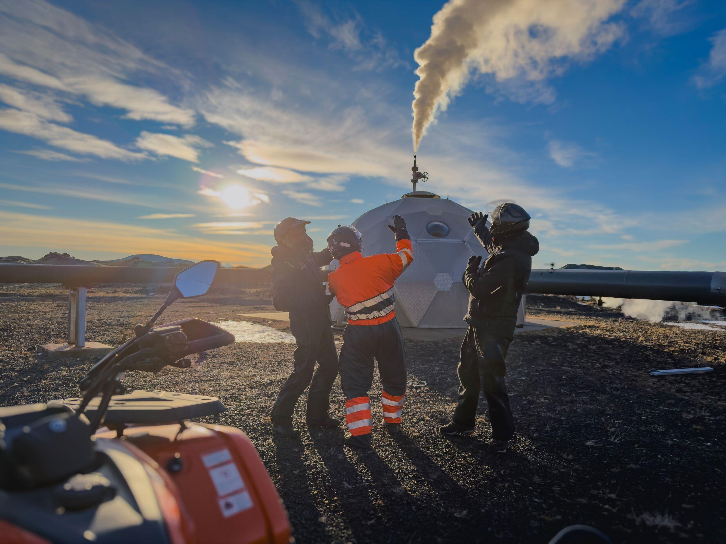 Three people stand near an ATV, a steaming geodesic dome, and industrial pipes in a sunny, rocky landscape.