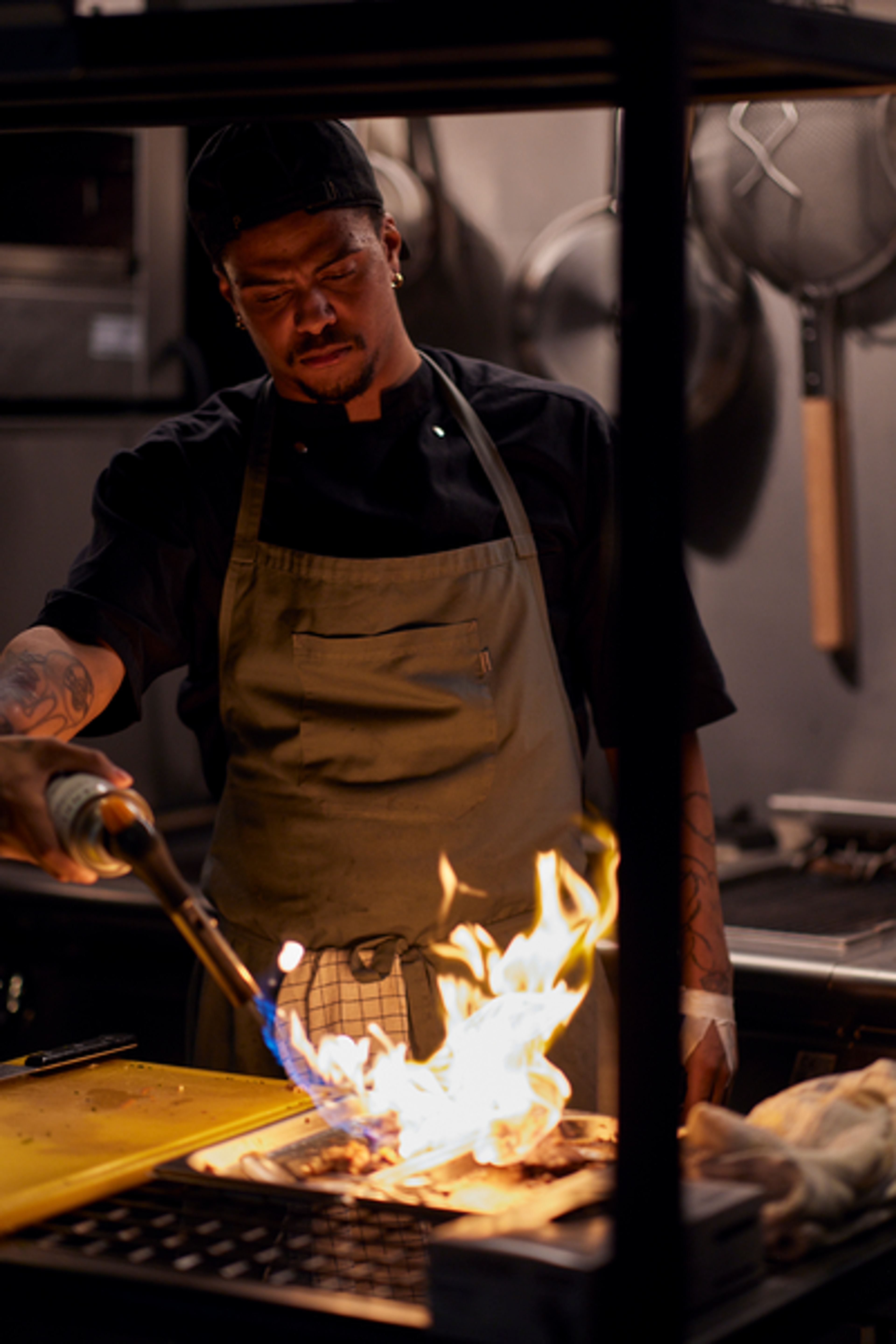 Chef using a blowtorch to cook food in a dimly lit kitchen, wearing a black hat and apron.