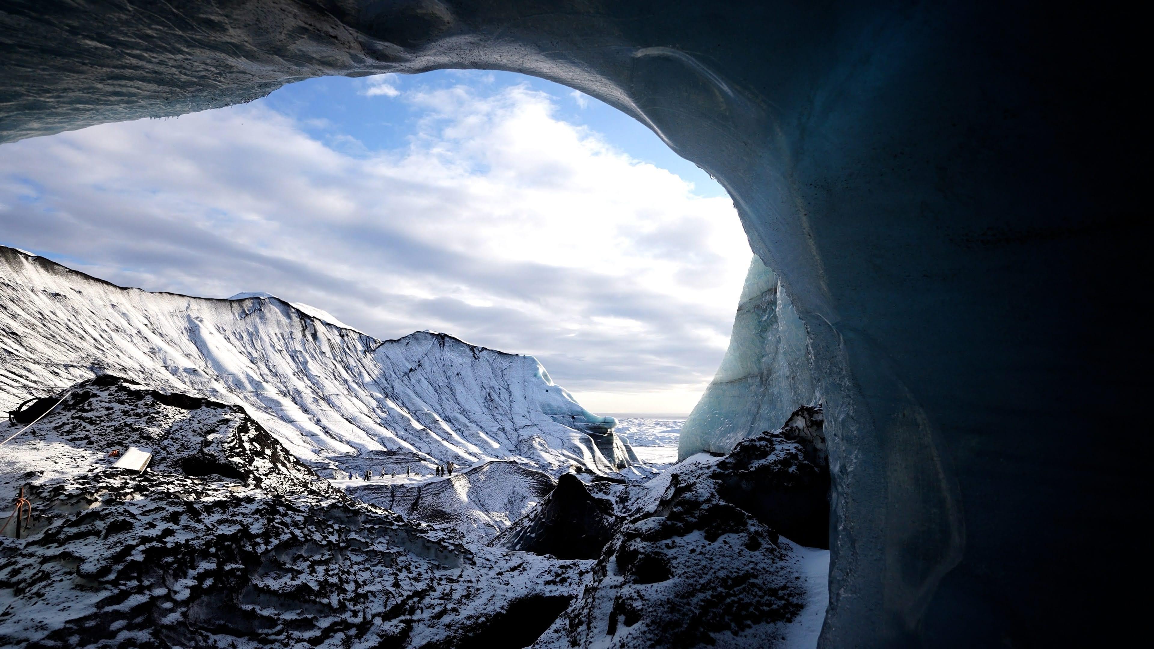 A dark blue ice cave opening frames a bright snowy mountain landscape under a cloudy sky.