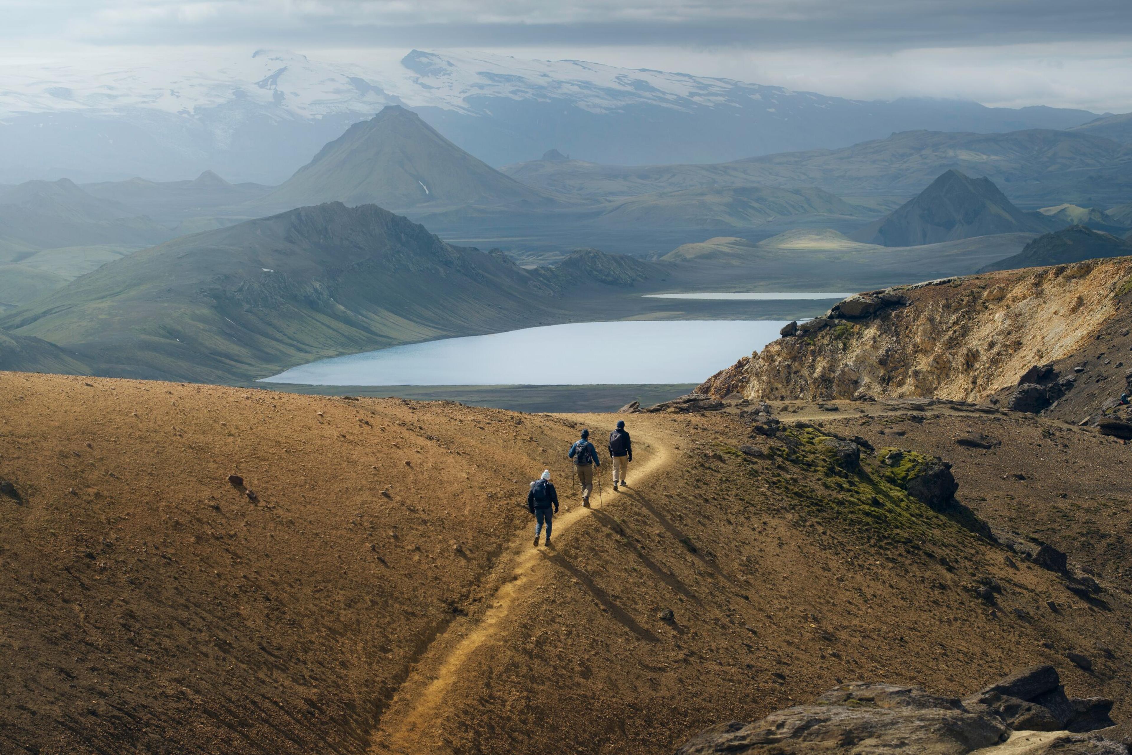 Hikers descending a trail on a rugged hillside with a panoramic view of a lake and mountainous landscape in the distance.