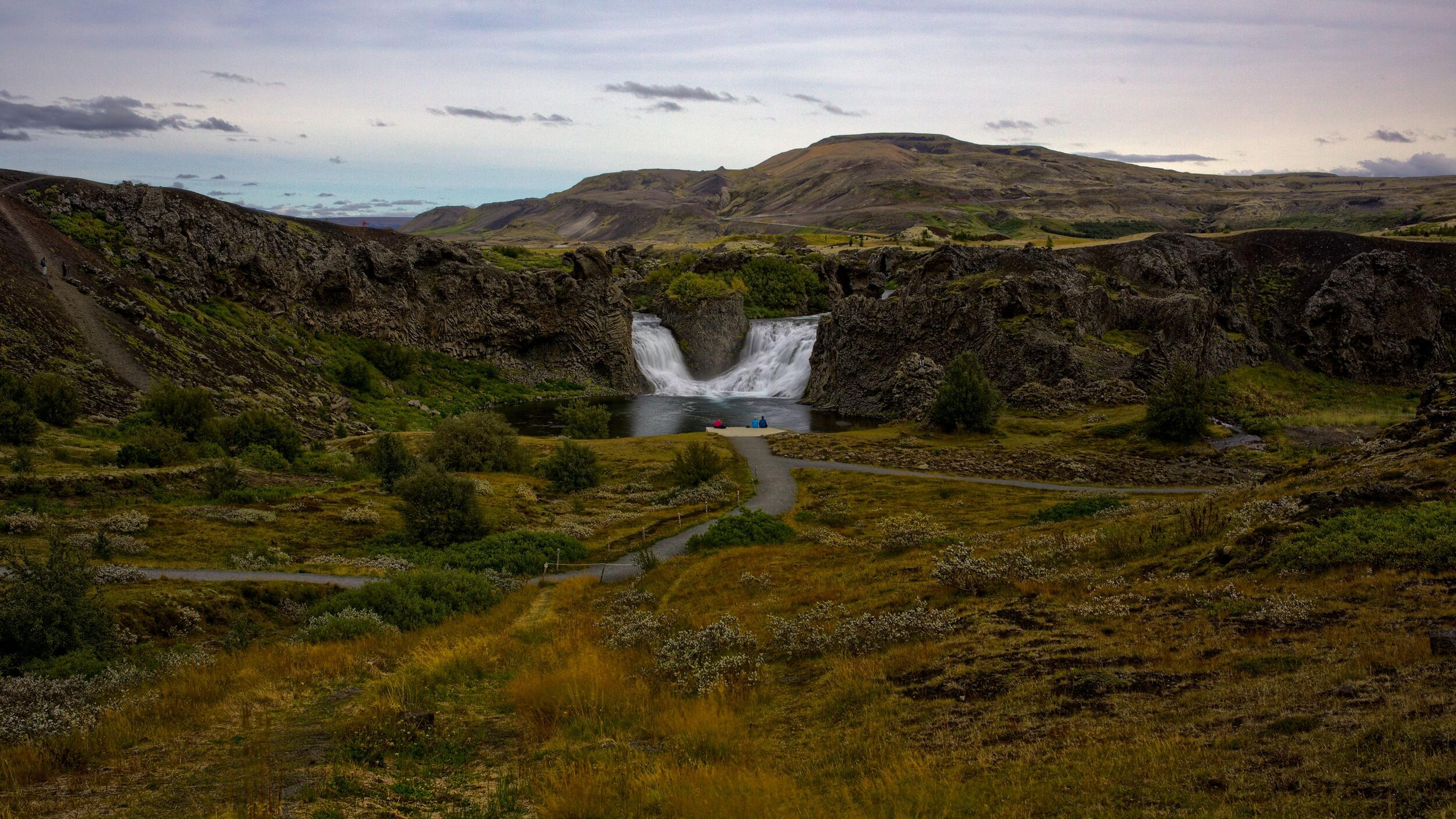 A waterfall cascades into a pool in a rocky, vegetated valley with a winding path.