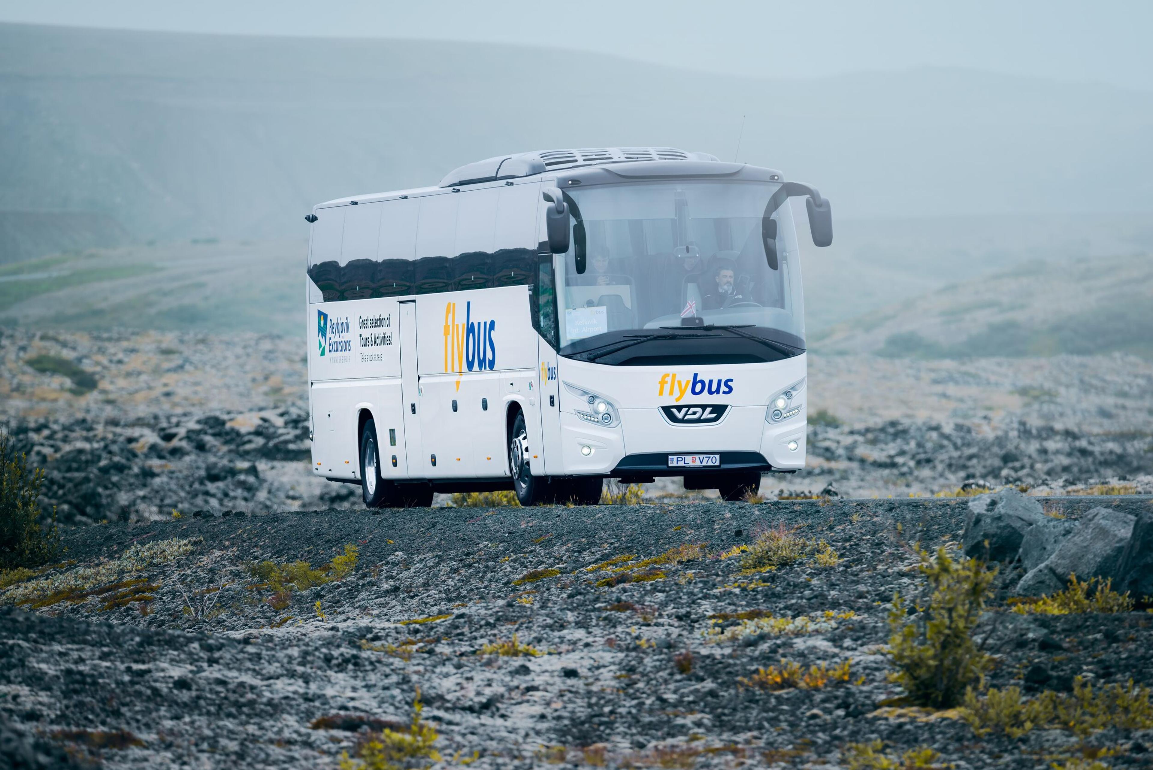 The Blue Lagoon shuttle bus travels through the terrain, with the shimmering lagoon visible in the background.