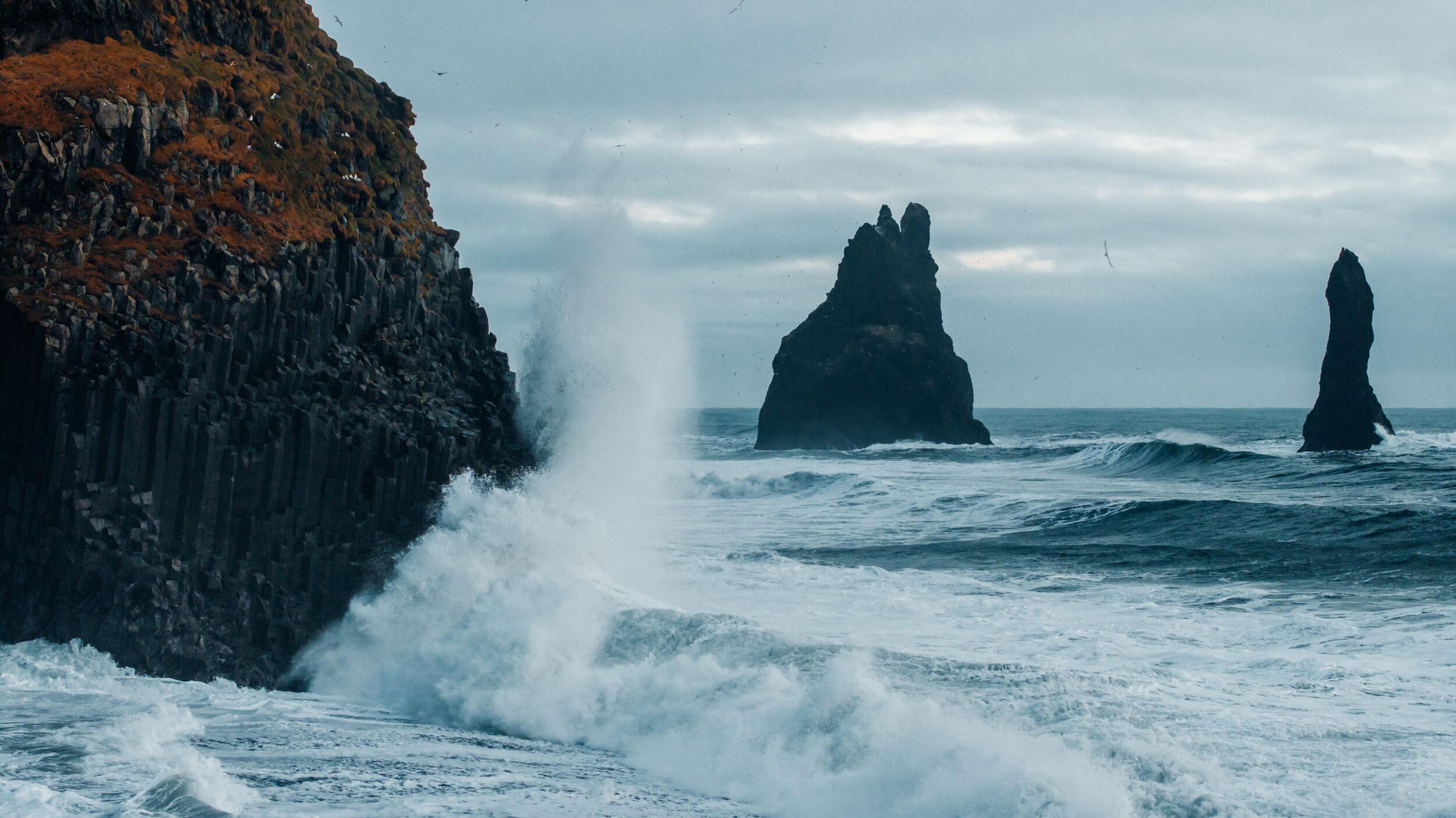 Waves crash on a dark columnar basalt cliff, with two sea stacks in the turbulent ocean under a cloudy sky.
