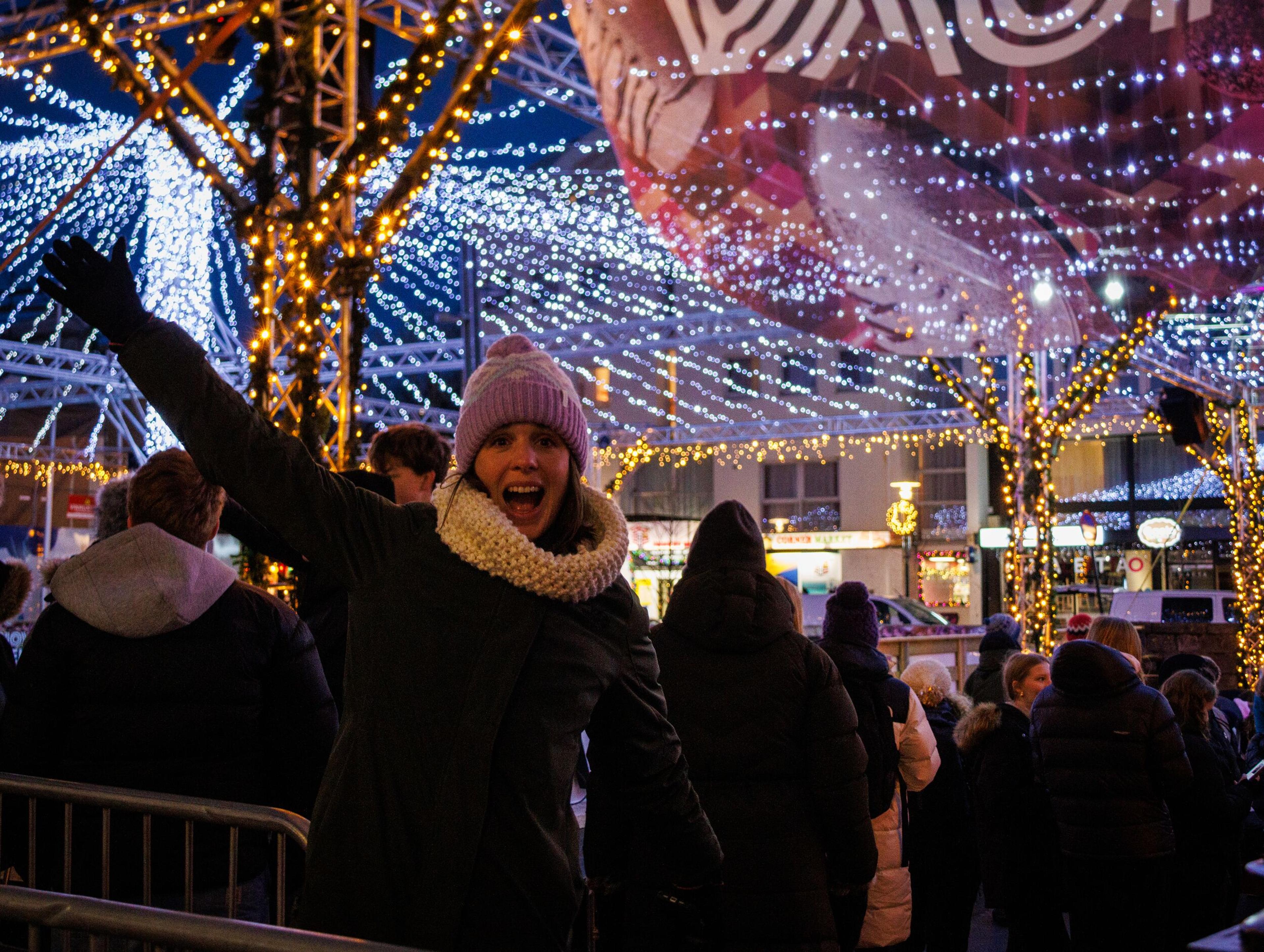 A smiling woman in a purple hat and white scarf raises her arm at a winter festival filled with sparkling lights.