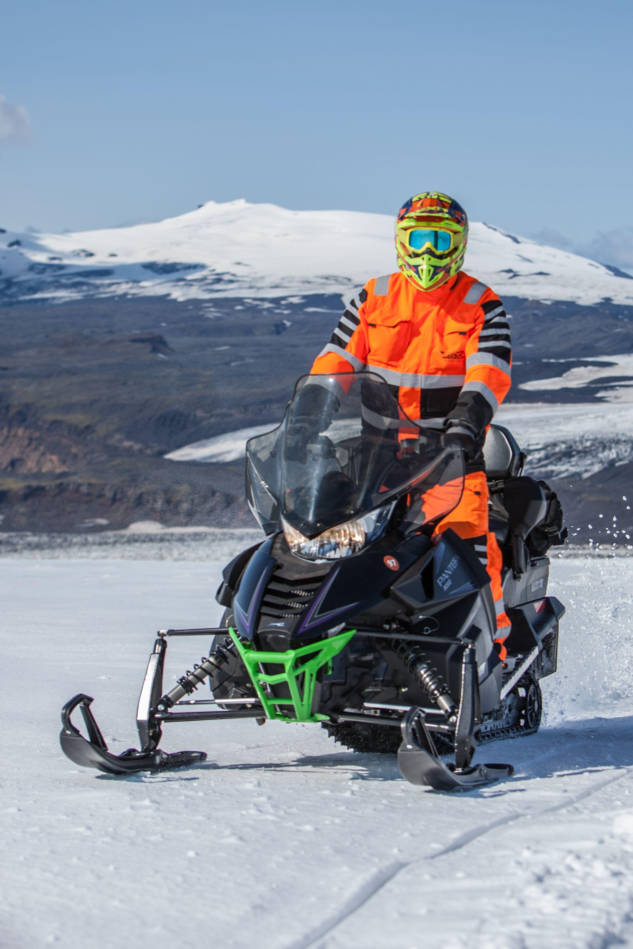 A person in bright orange gear and a helmet riding a snowmobile across an Icelandic glacier, with snow-covered mountains in the background.