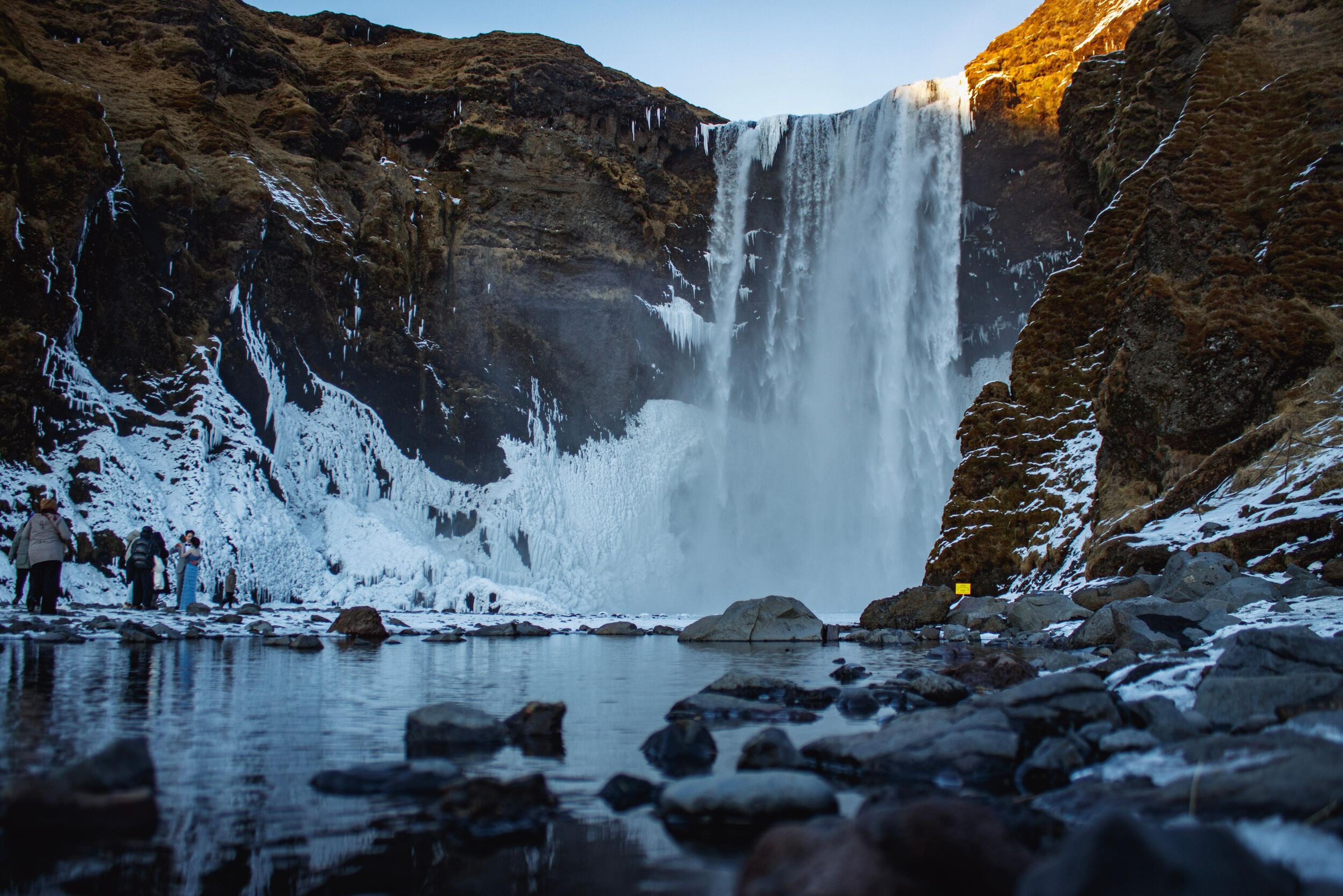 A large waterfall plunges between snow-covered cliffs into a rocky pool, with several people visible at its base.
