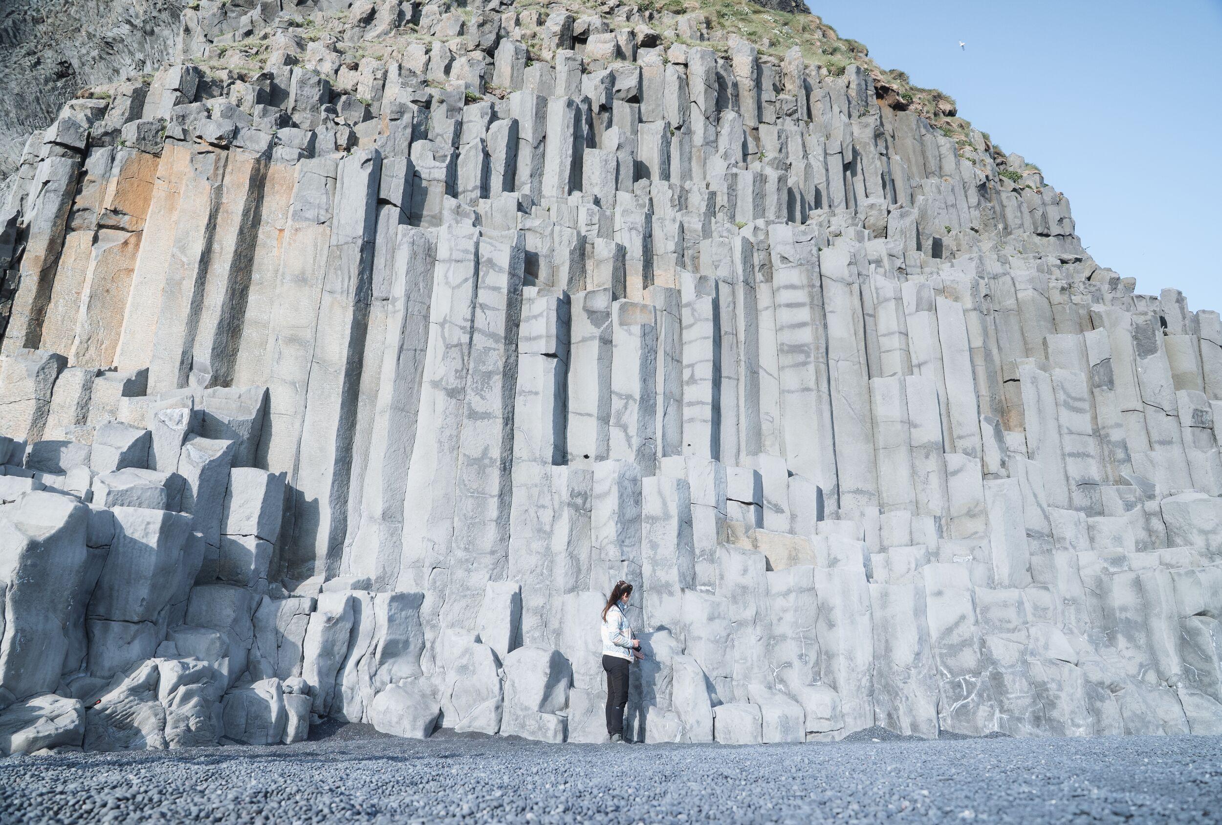 A person stands on a dark pebbly beach at the base of a towering cliff of grey basalt columns.