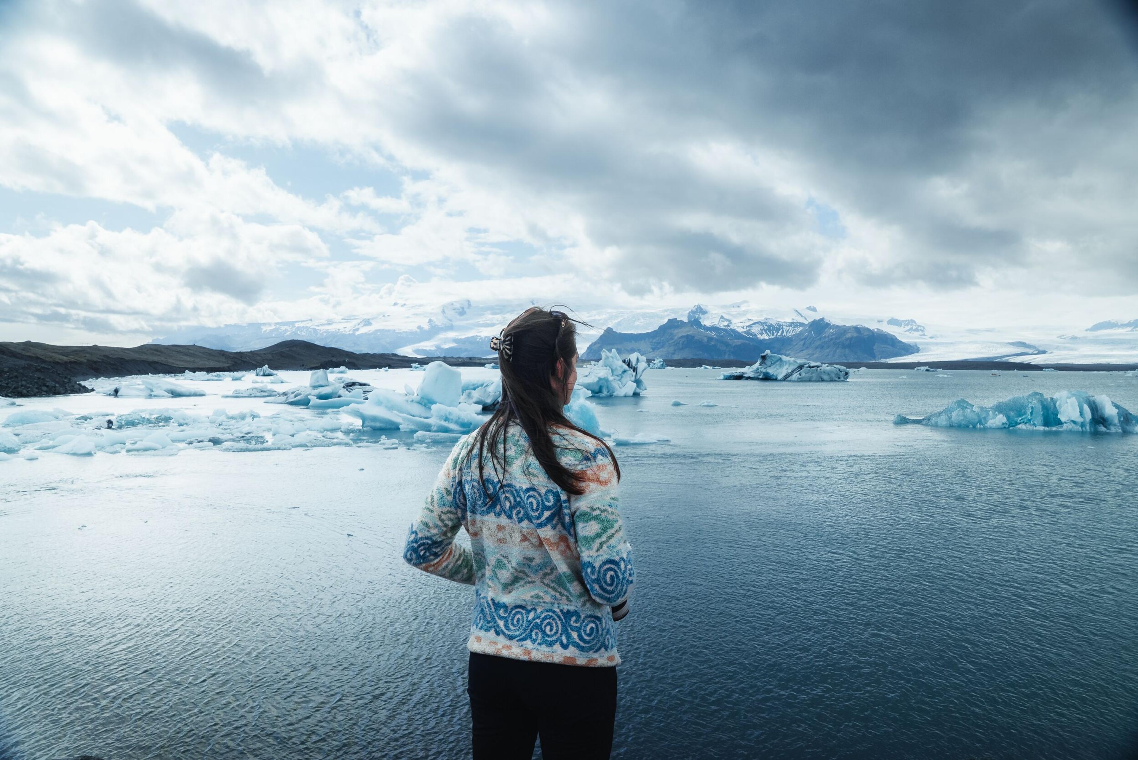 Person from behind views a glacial lagoon with icebergs, mountains, and a cloudy sky.