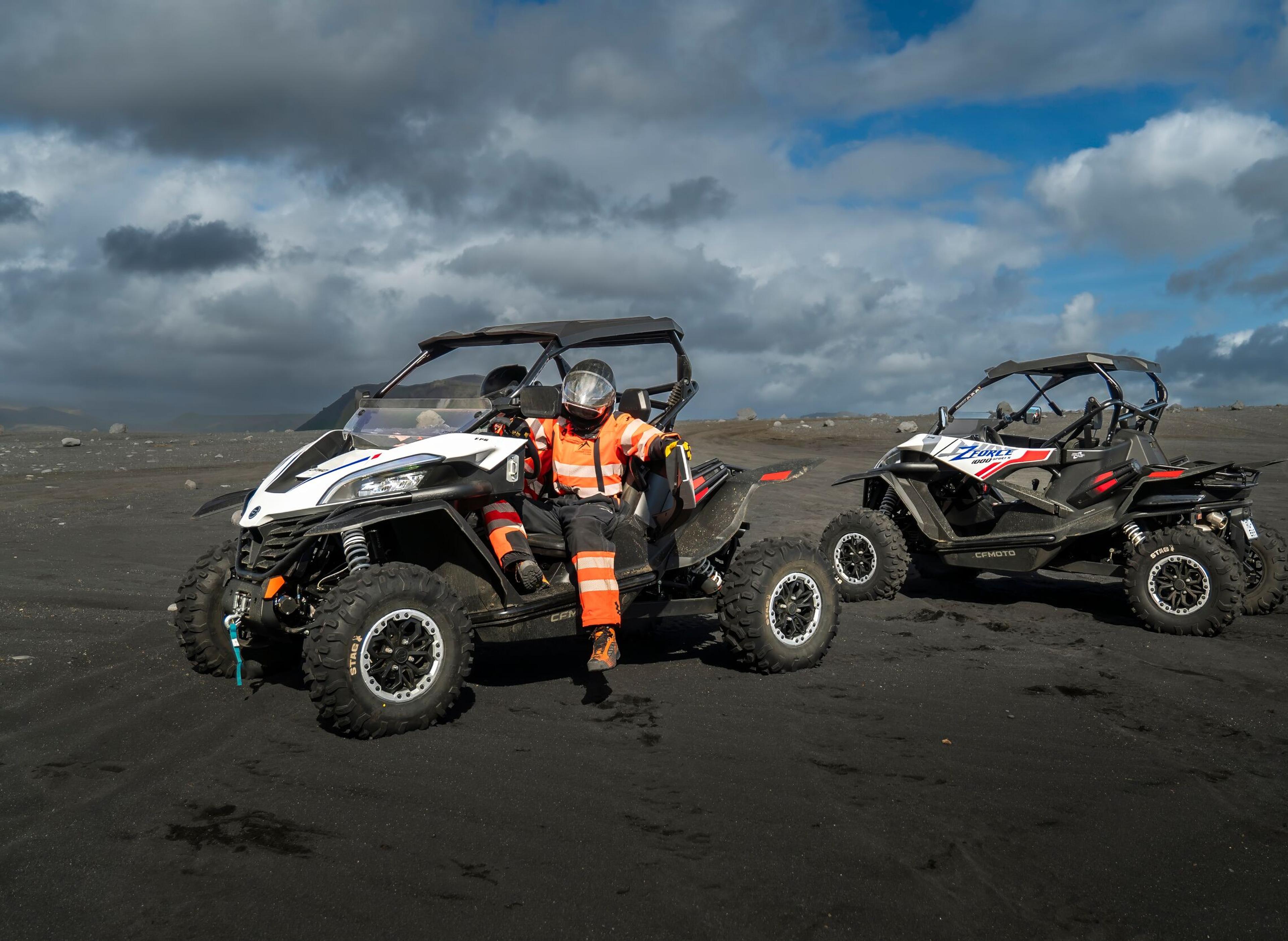 Person in orange gear sitting on an off-road vehicle with another similar vehicle nearby on a black sand landscape in Iceland.