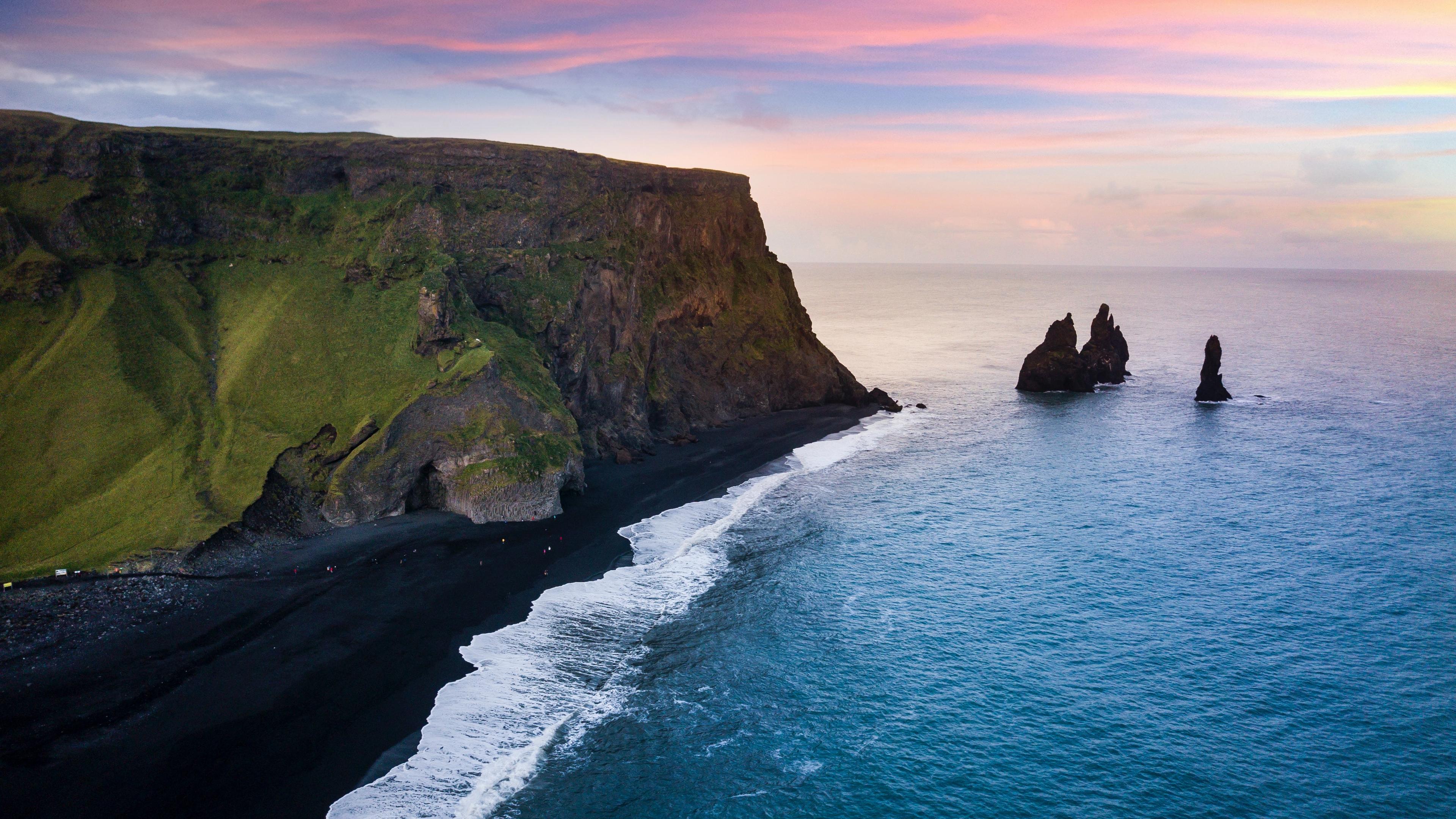 Aerial perspective of Reynisfjara beach bathed in a soft pink hue from sunset, highlighting the vast black sands, jagged coastline, and distant sea stacks, creating a surreal and dreamy landscape in Iceland.