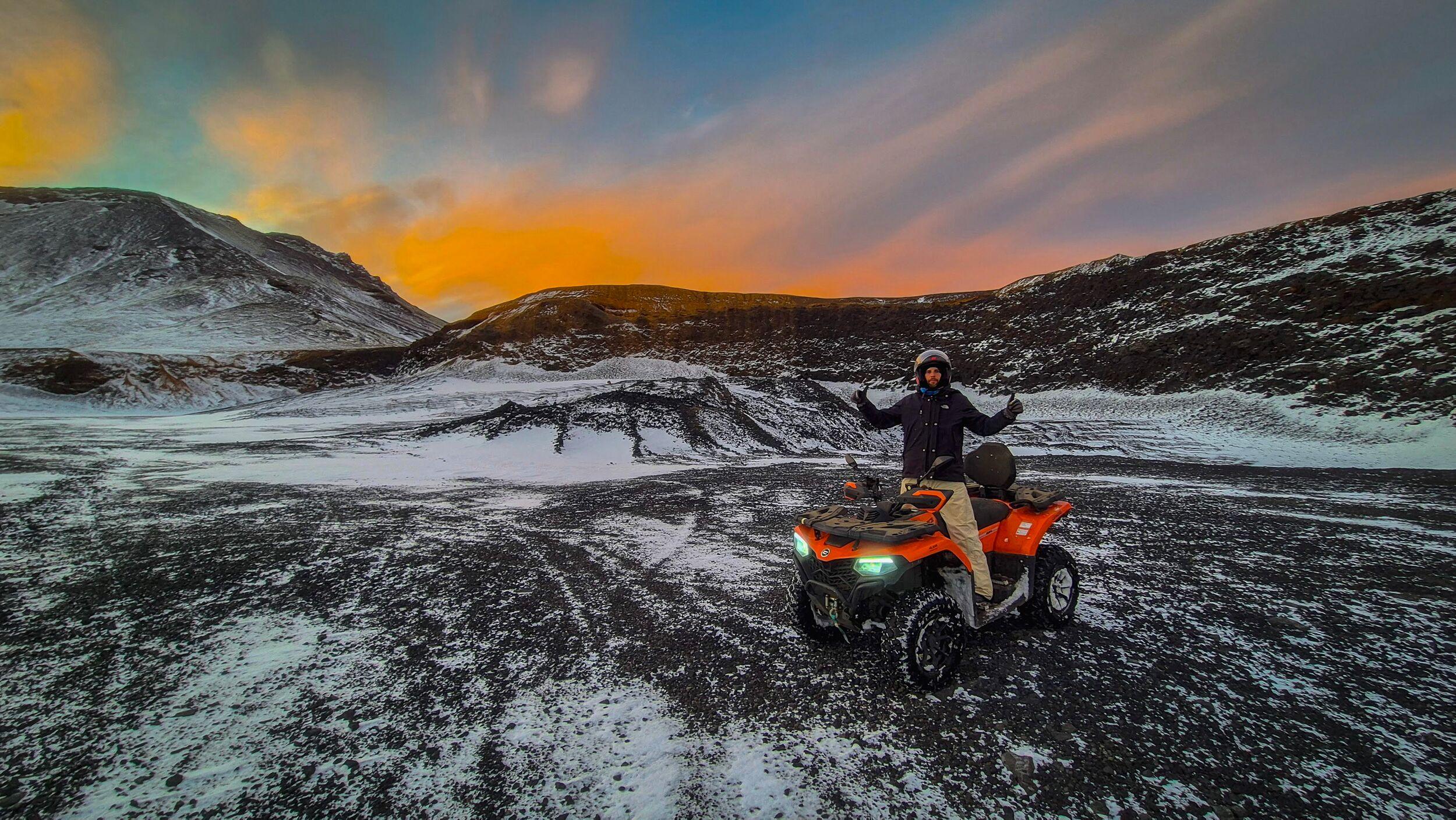Man on an orange ATV in a snowy volcanic landscape at sunset.