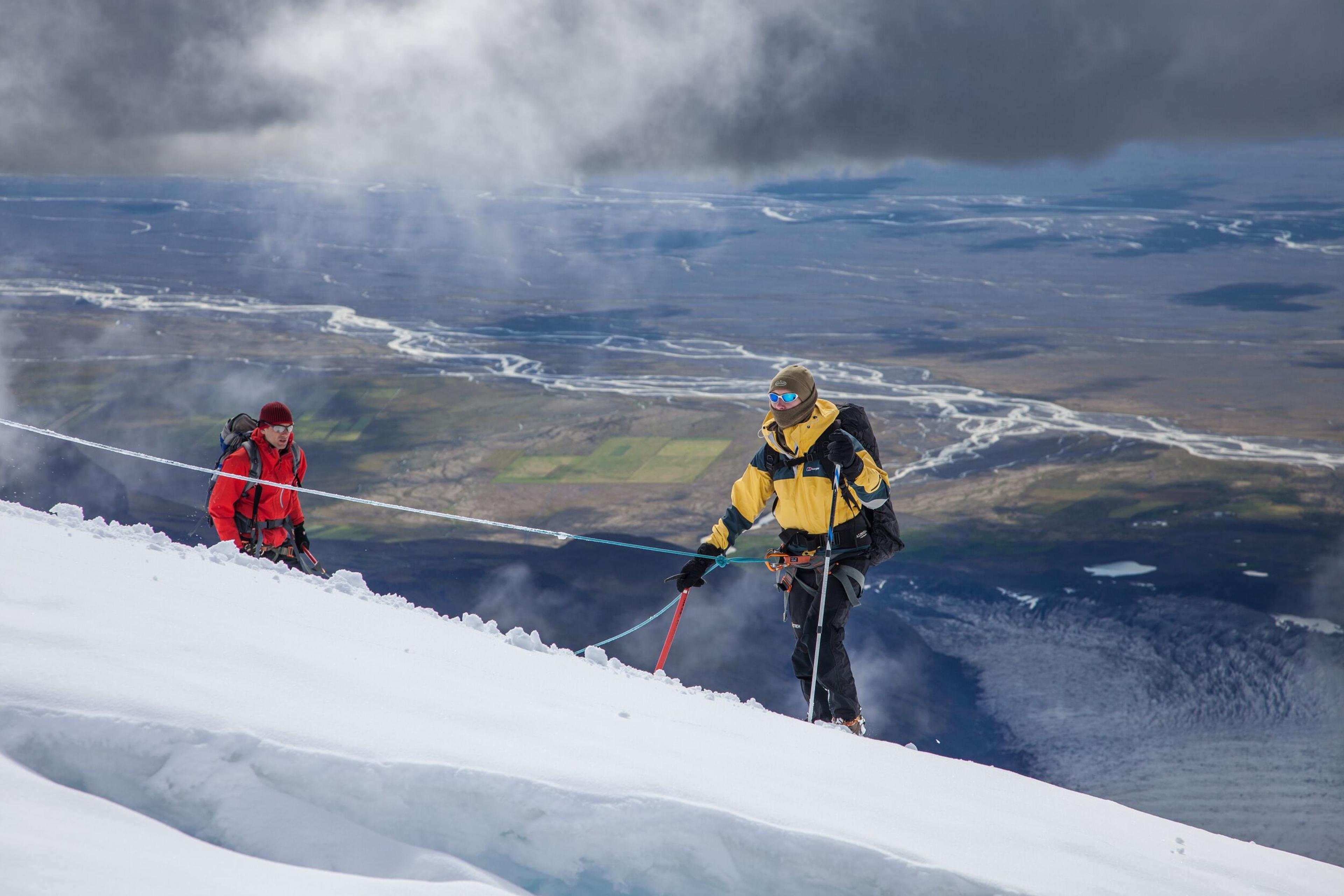 Two roped mountaineers ascend a snowy slope overlooking a vast valley with rivers.