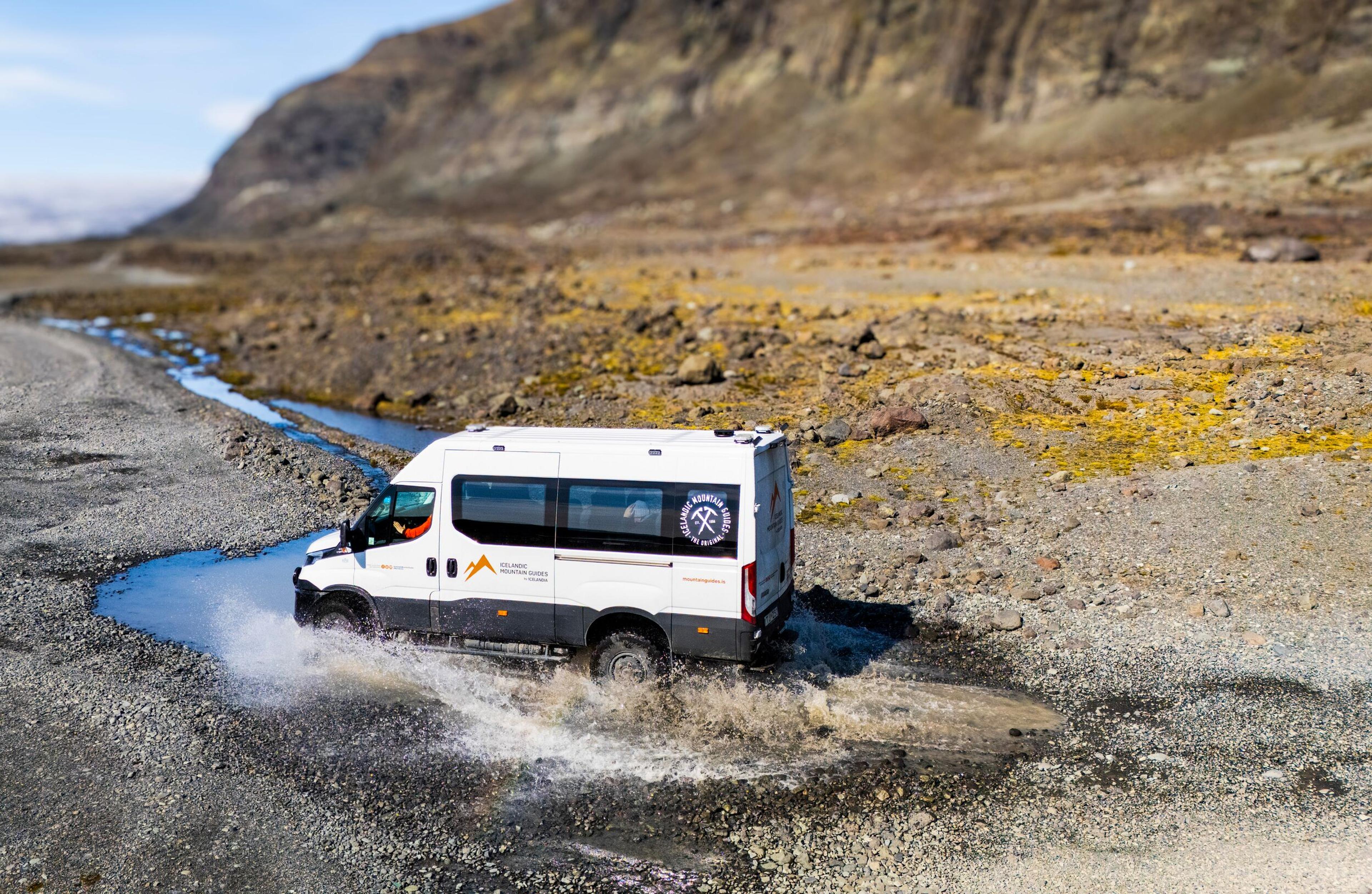 A white off-road van splashes through water on a rocky road in a barren landscape.