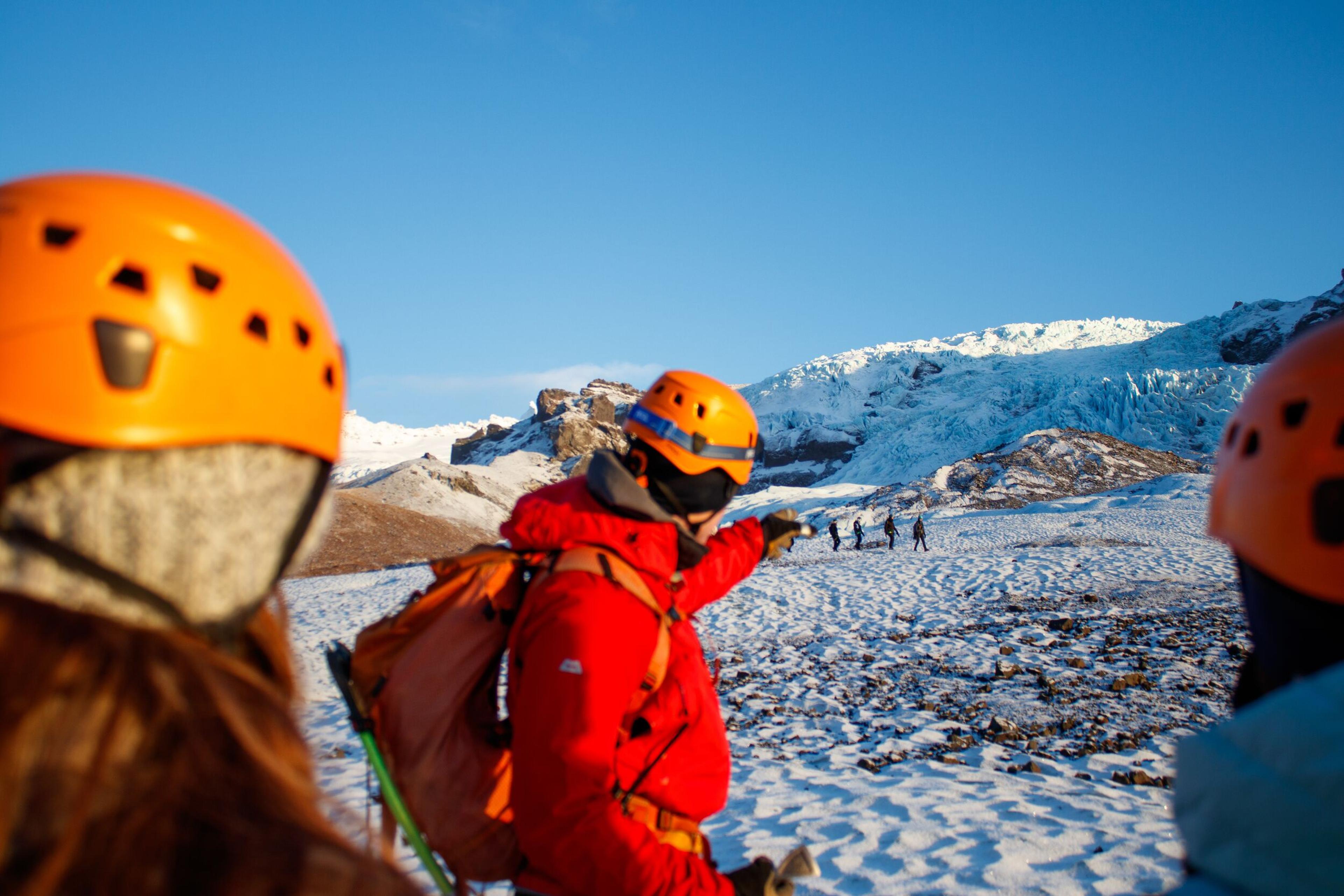 A group of hikers in orange helmets on a snowy glacier, one pointing towards distant figures and an ice-covered mountain.