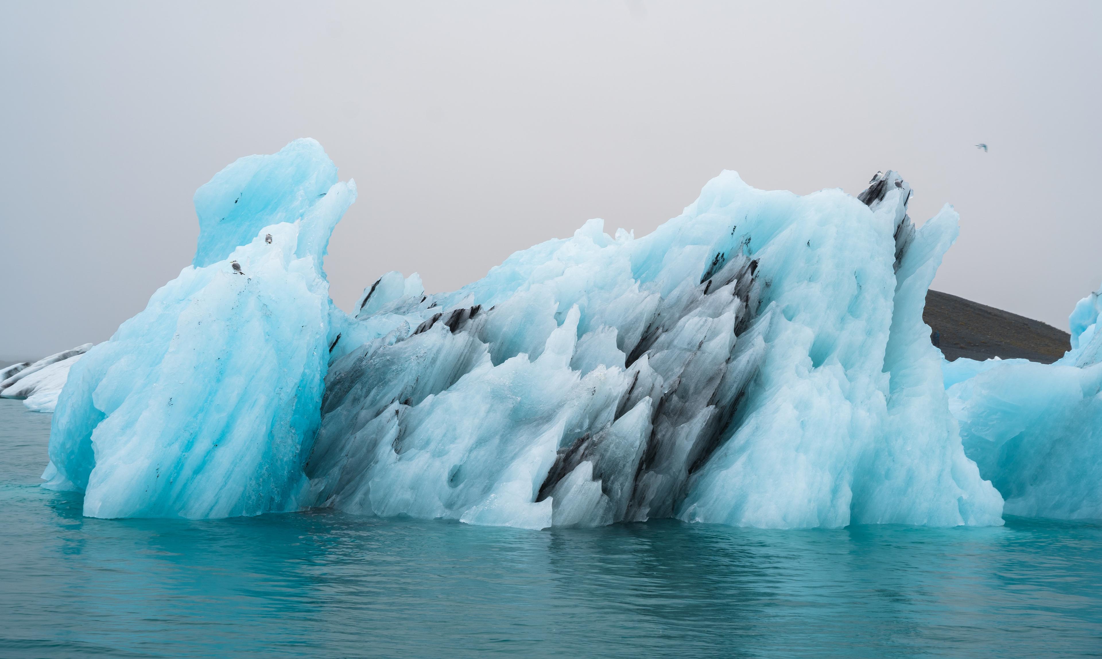 A craggy ice berg floating on the lake, displaying all hues of blue and enriched with black ash layers