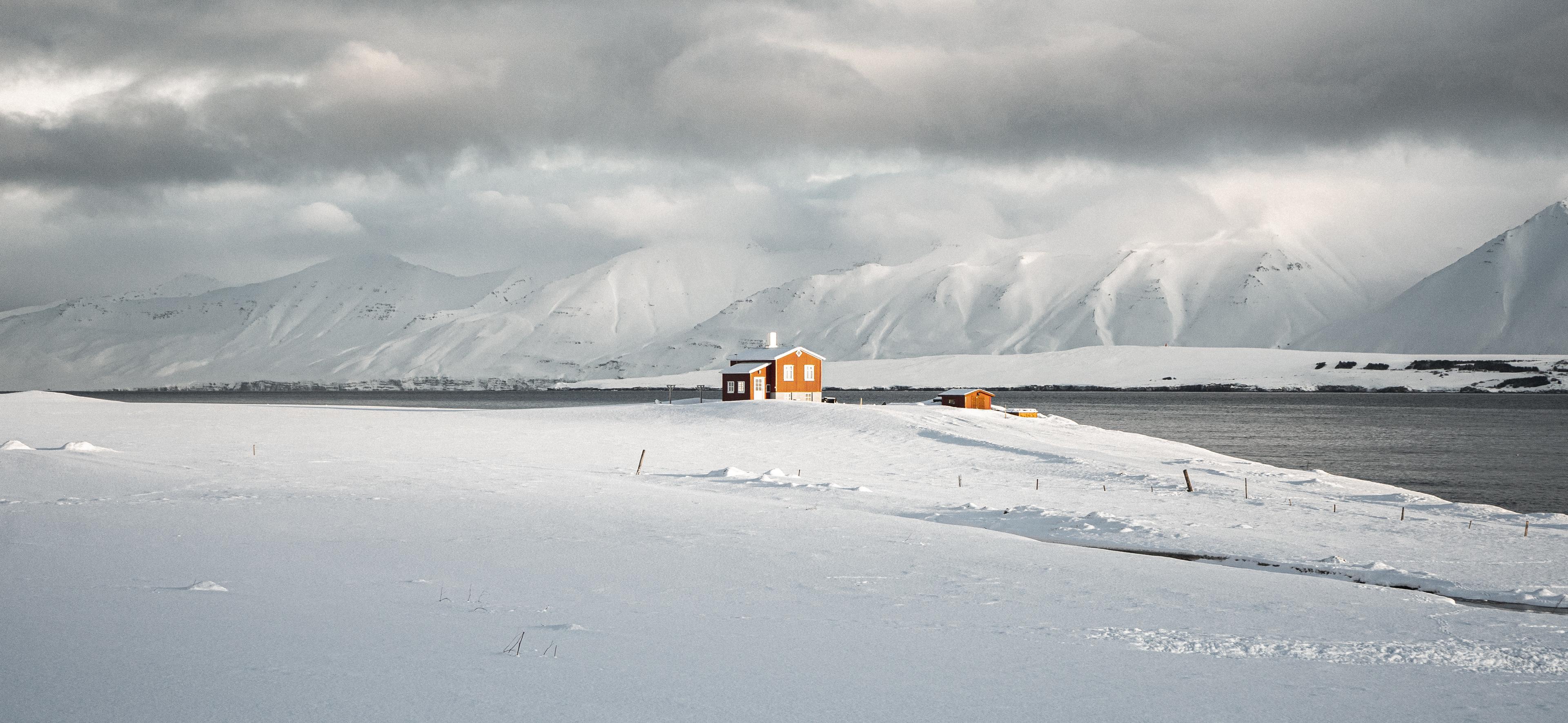 A small orange house stands out against a vast snowy landscape with a backdrop of snow-covered mountains under a cloudy sky.