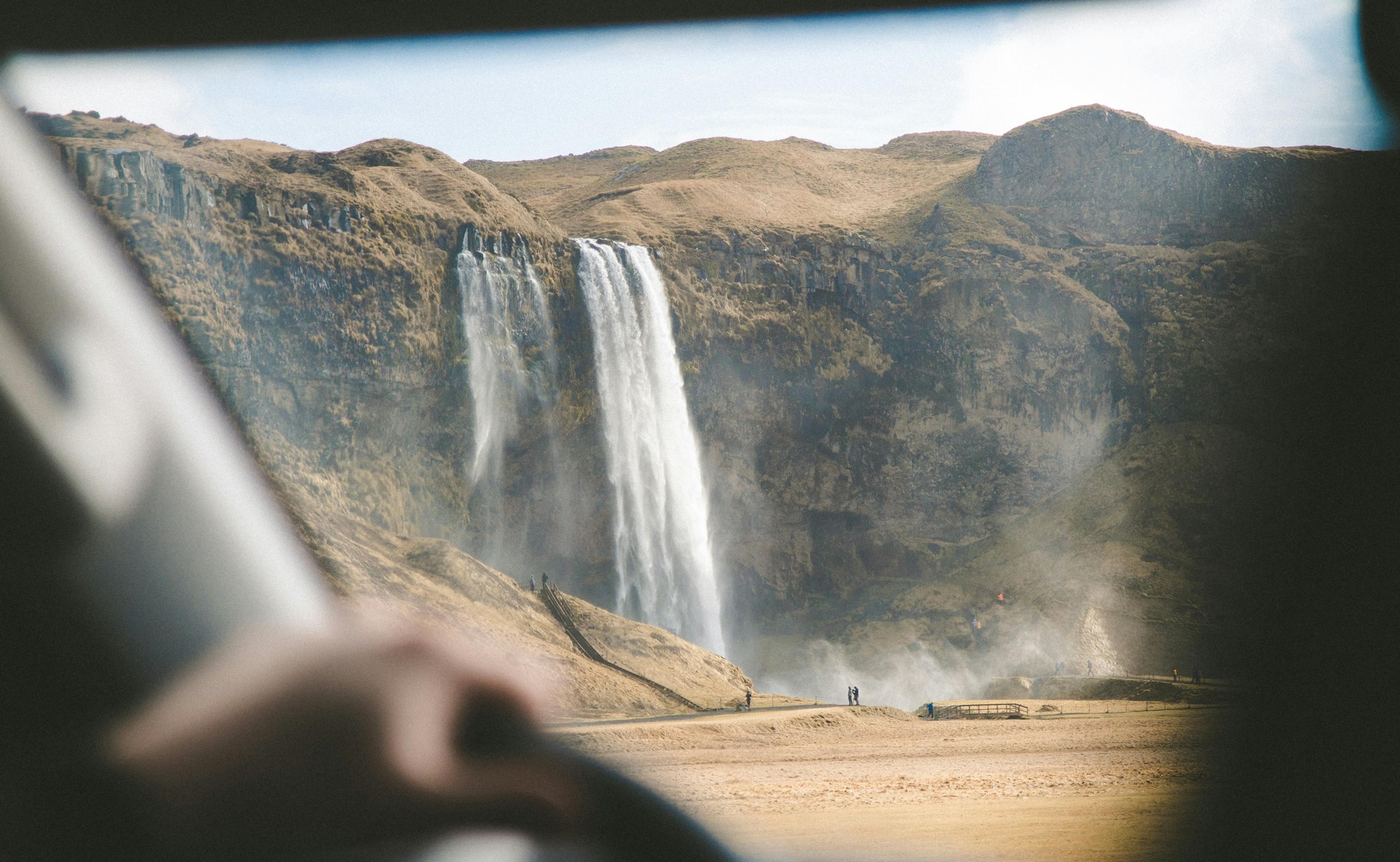 View from inside a car showing a person looking at a large waterfall cascading over a cliff with mist rising from its base.