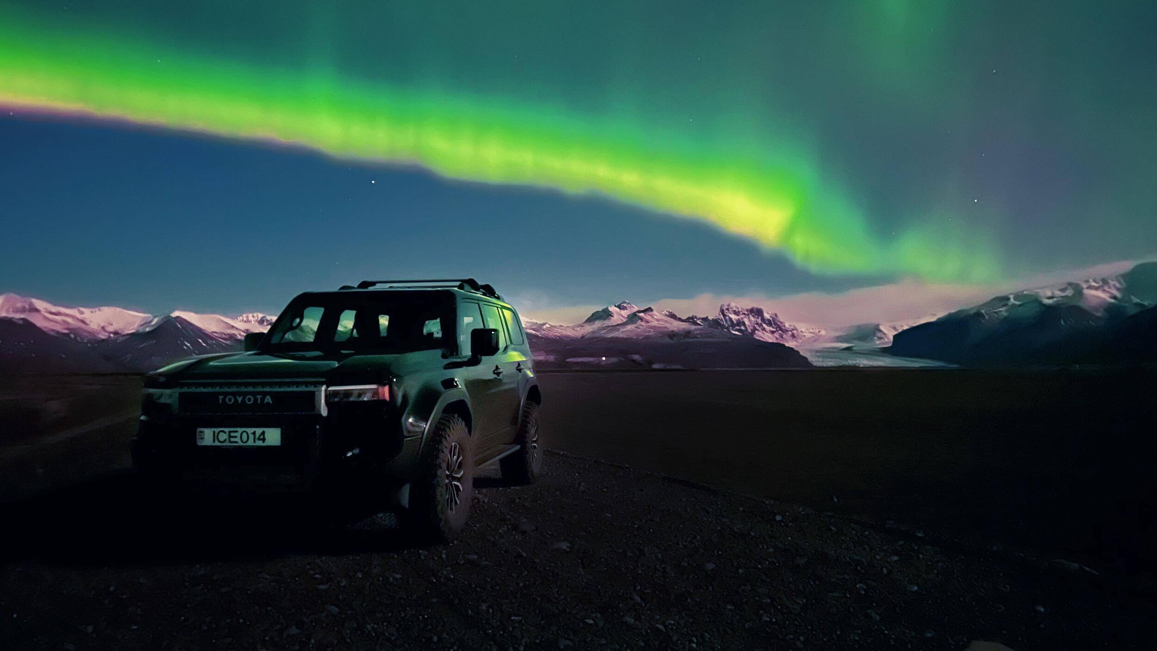 Dark Toyota SUV under vibrant green Northern Lights with snow-capped mountains.
