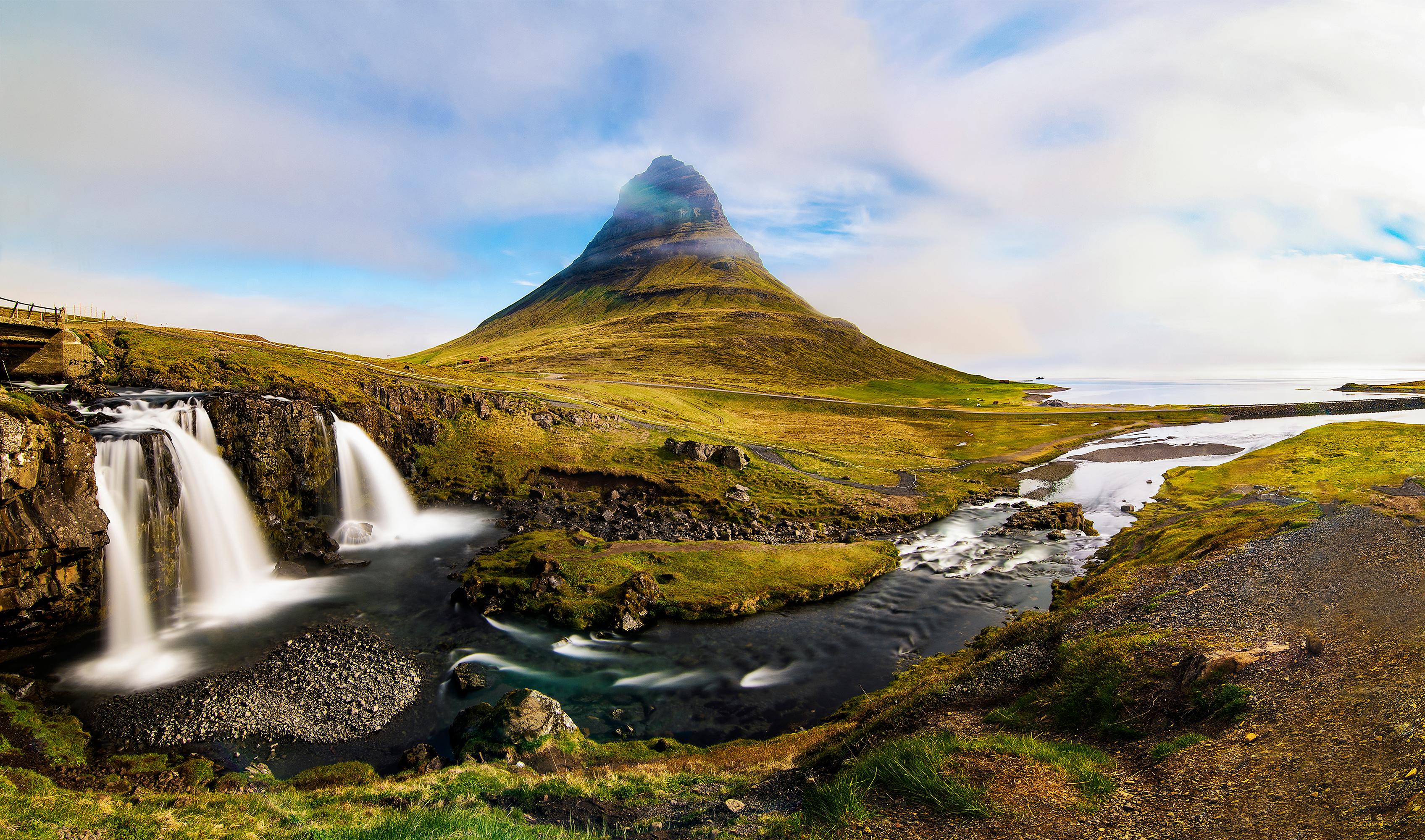 Kirkjufell mountain partially shrouded in mist, overlooking Kirkjufellsfoss waterfalls with smooth, flowing water, and green landscape.