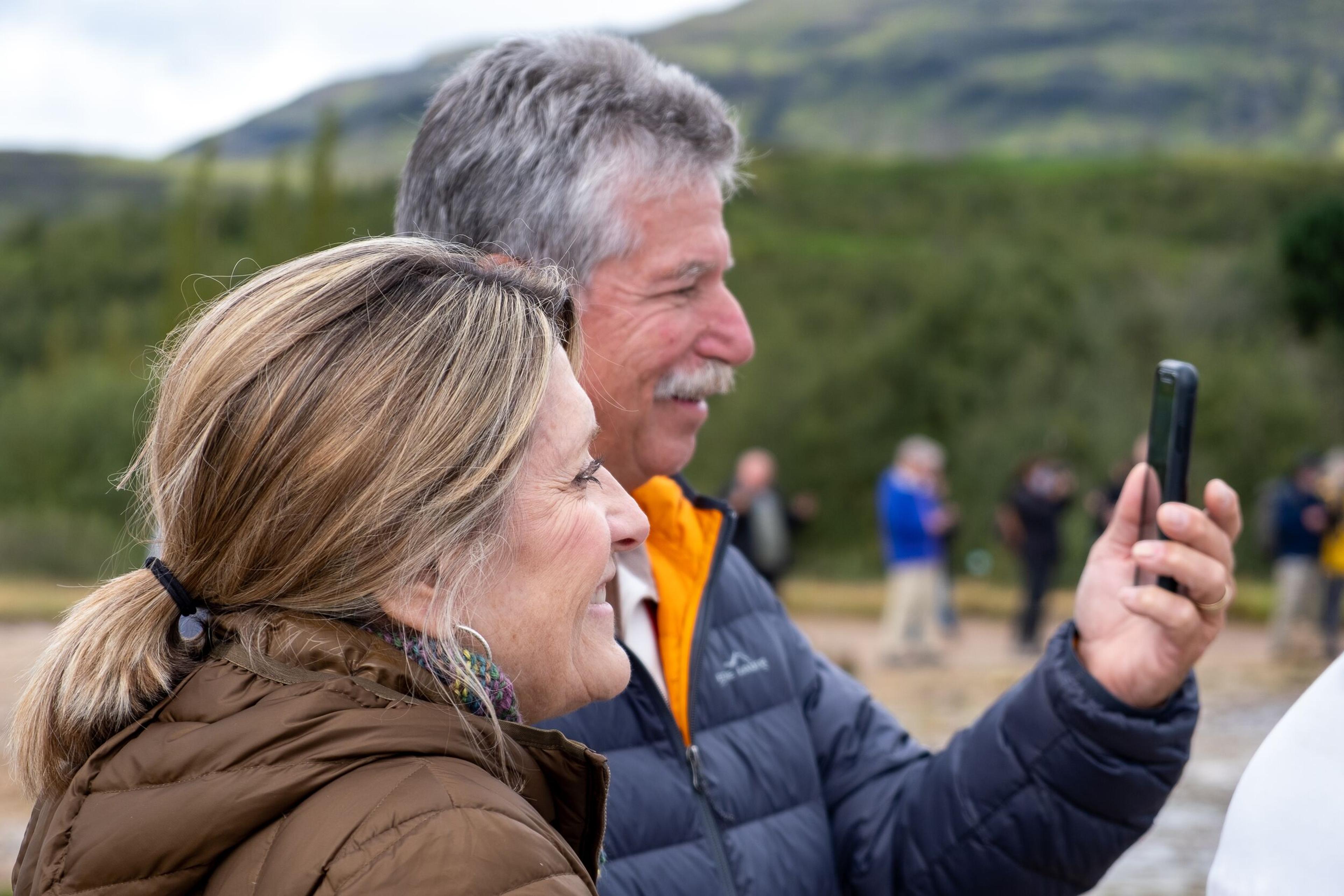 Two adults are smiling and taking a selfie with a smartphone, with other tourists and a green landscape in the background.
