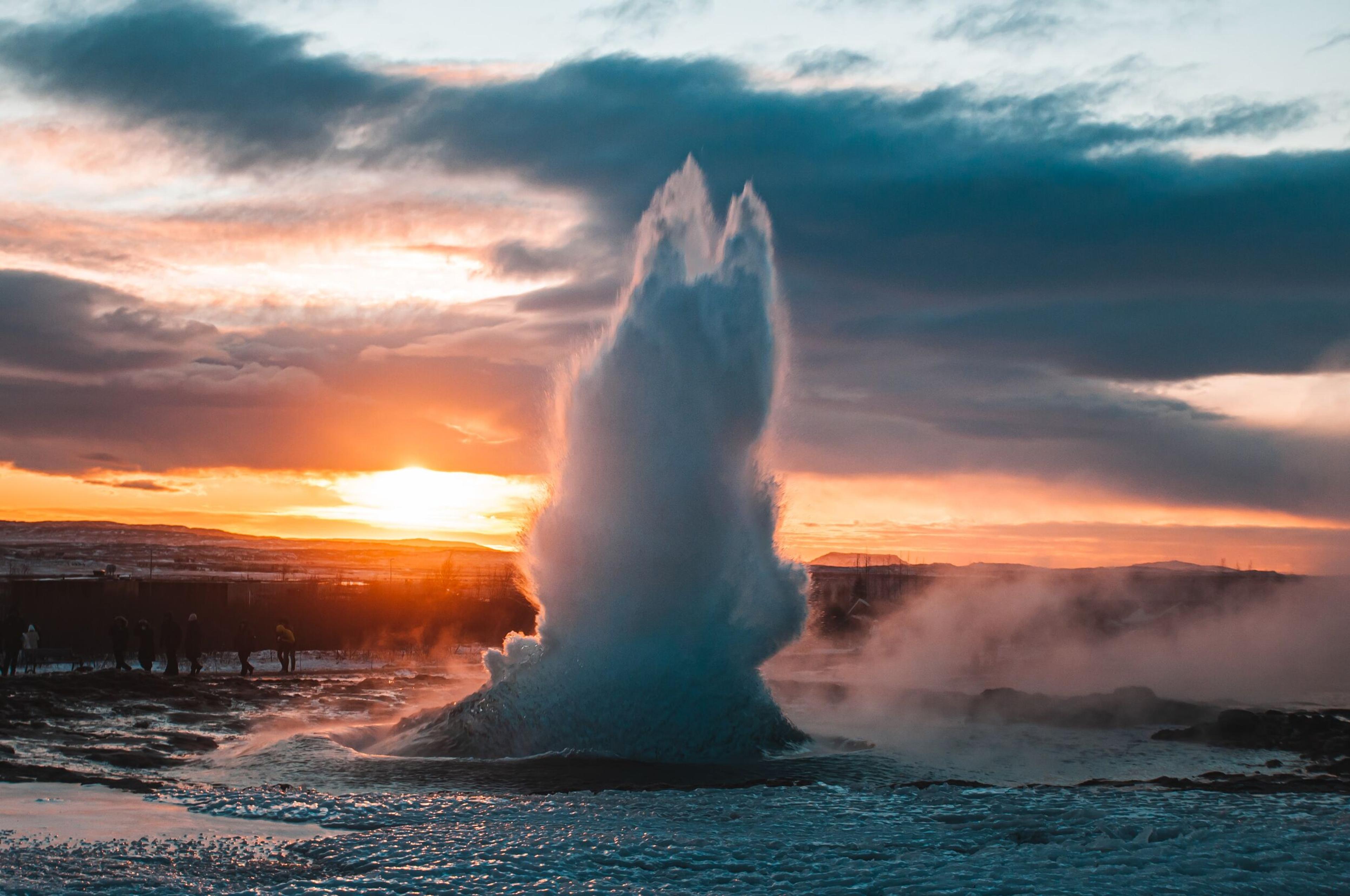 A geyser erupts with a powerful spray of water and steam at sunset, creating a dramatic scene against the orange and pink hues of the sky. People in the background observe the natural phenomenon.