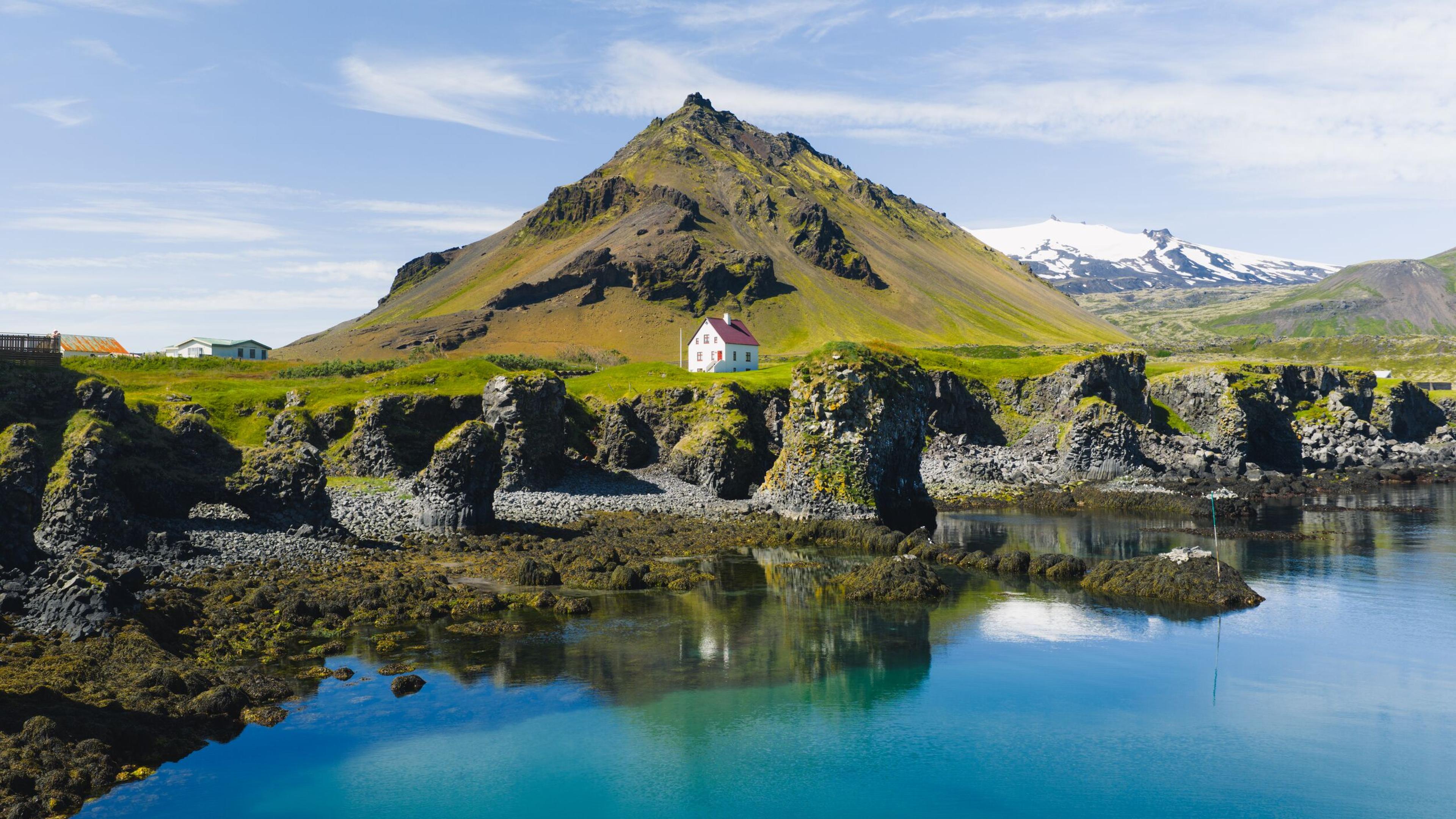 A small white house with a red roof on a green hill overlooks a rocky bay, with a dark mountain behind it and snowy peaks in the distance.