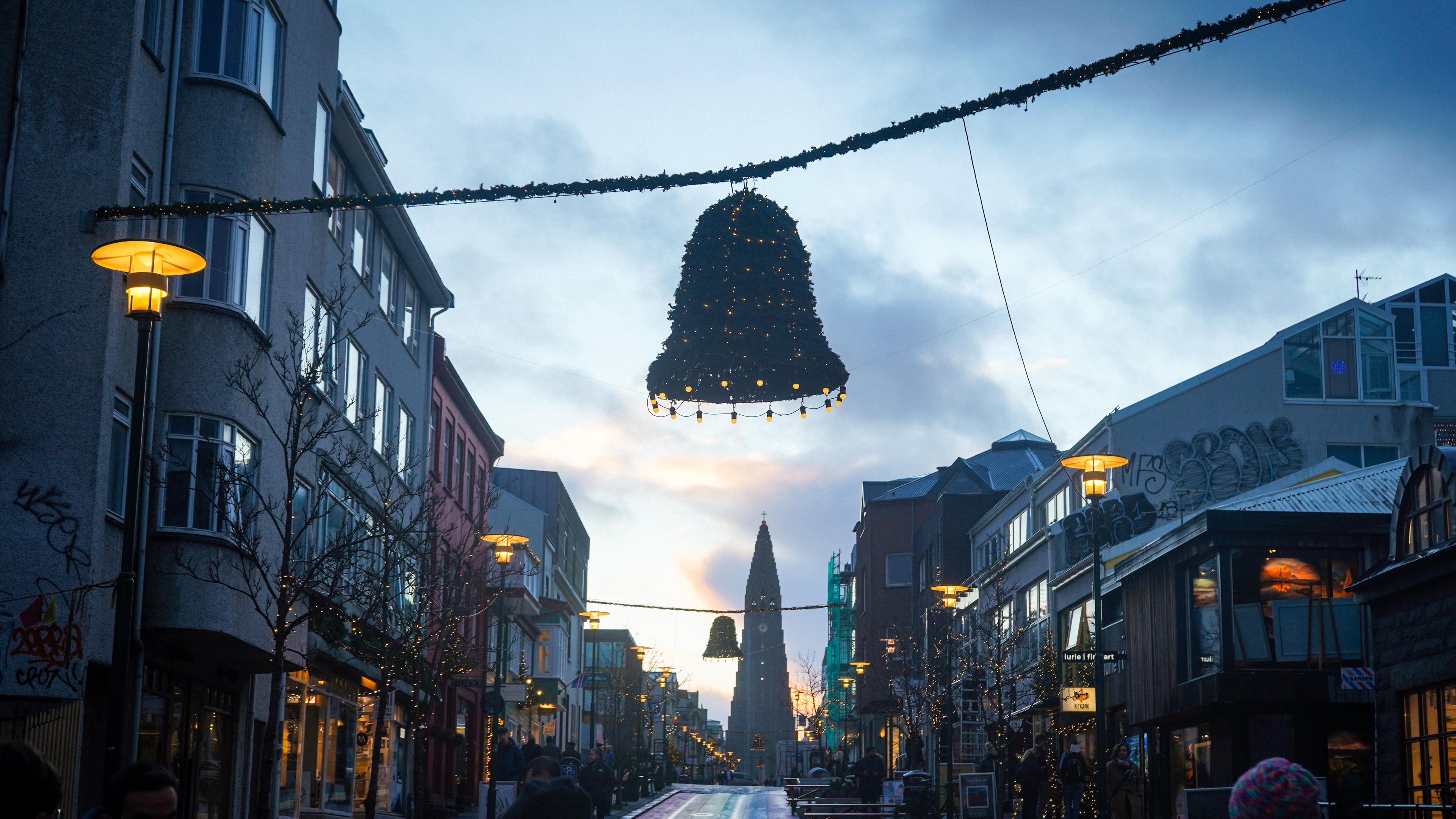 A picturesque view of a bustling street at dusk with buildings on either side, festive lights strung across the street, and a large Christmas tree-shaped light decoration hanging above