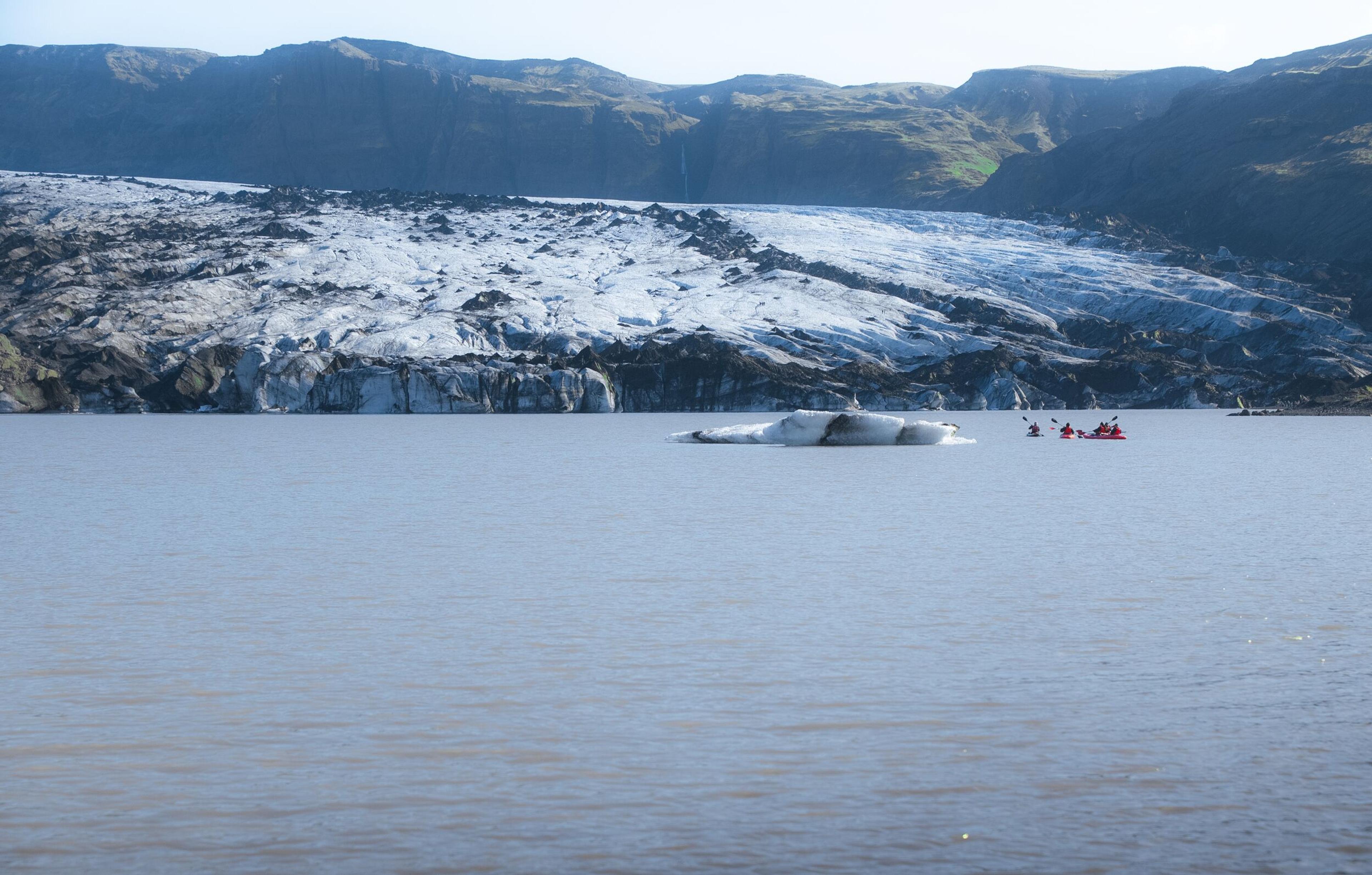 Kayakers paddle by an iceberg in a glacial lake, with a glacier and mountains behind.
