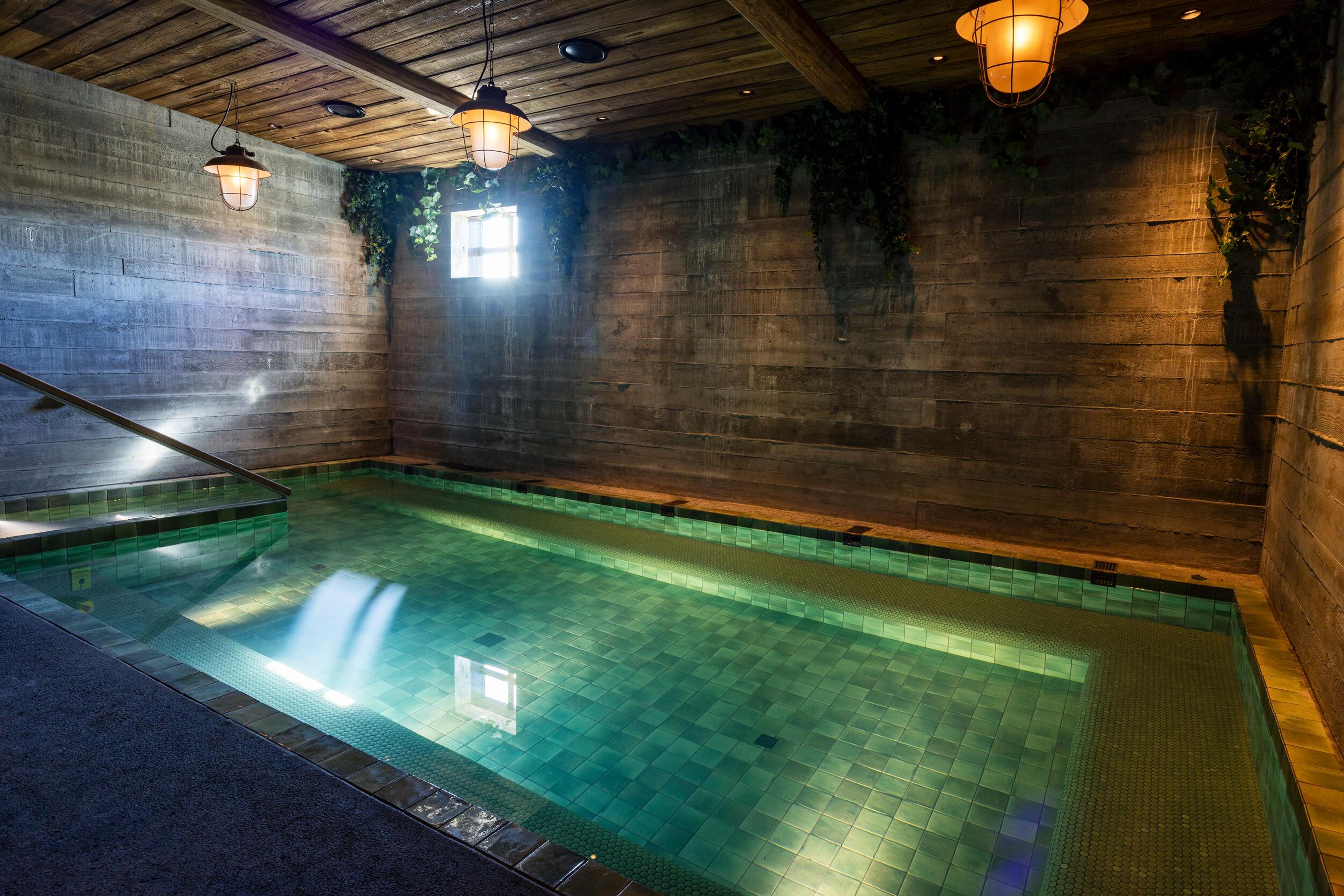 A rustic indoor pool with green tiles, dark textured walls, and a wooden ceiling. Light from a small window reflects on the water.