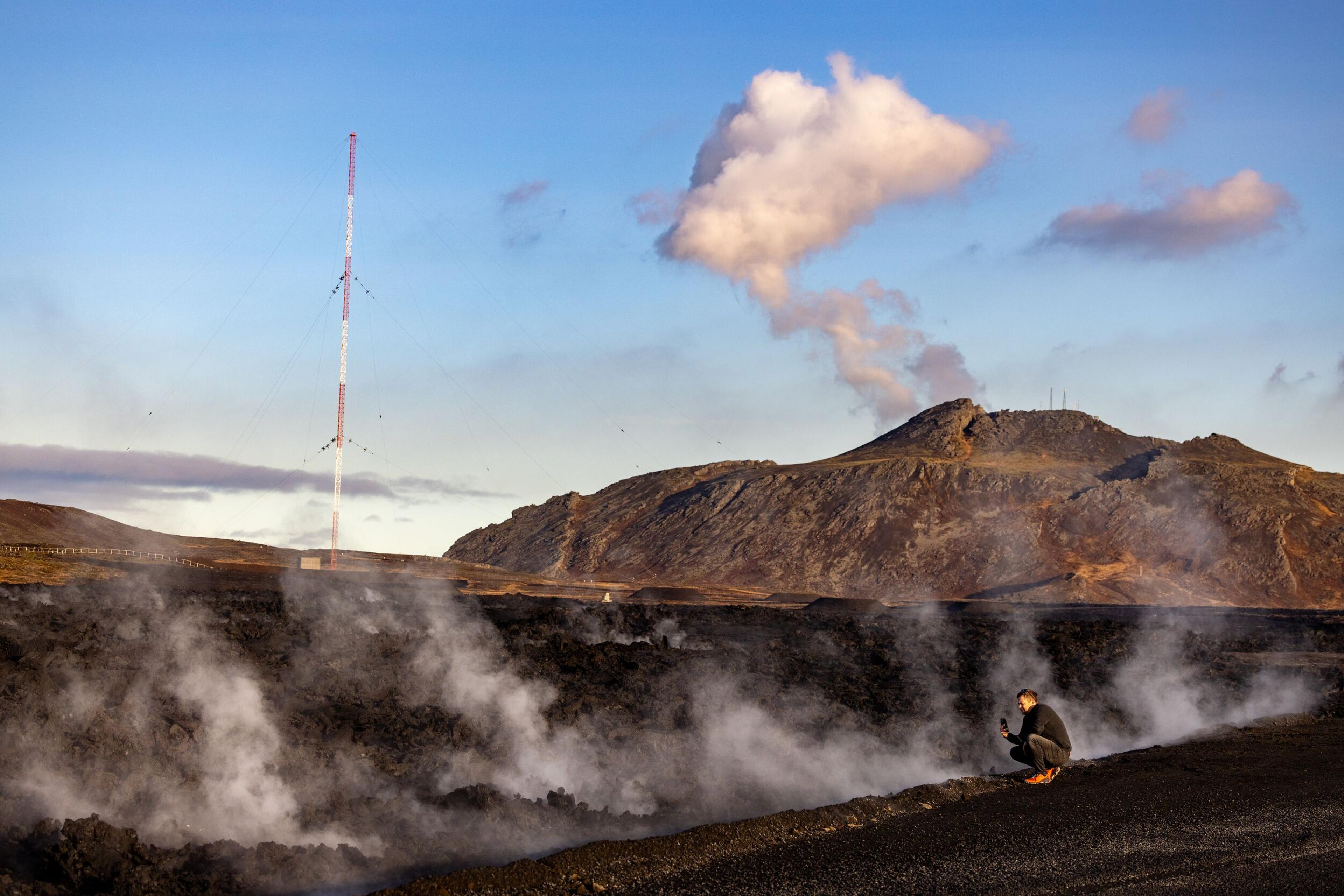 A person observes steaming lava fields near a volcanic site, with a plume of smoke rising from the mountain and a tall communication tower in the background under a blue sky.