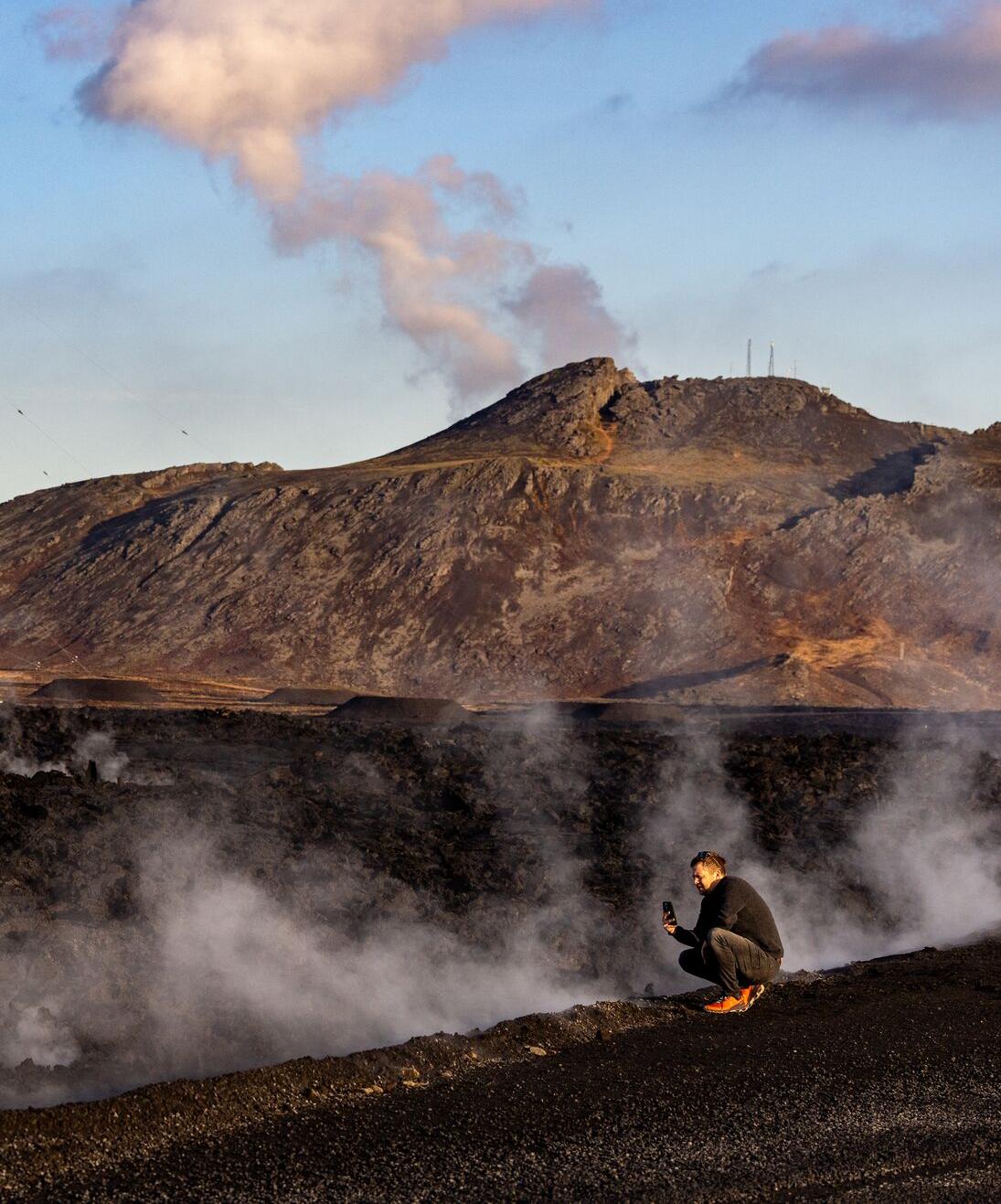 A person kneeling on a volcanic landscape surrounded by steam, with a smoking volcano and a clear blue sky in the background.
