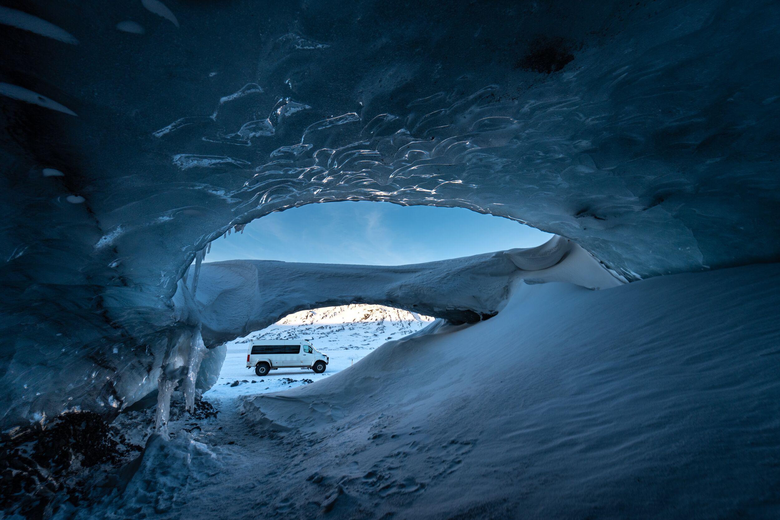 A large white off-road vehicle visible through the opening of an ice cave in Iceland, with icy formations, snow-covered landscape, and clear skies in the background.