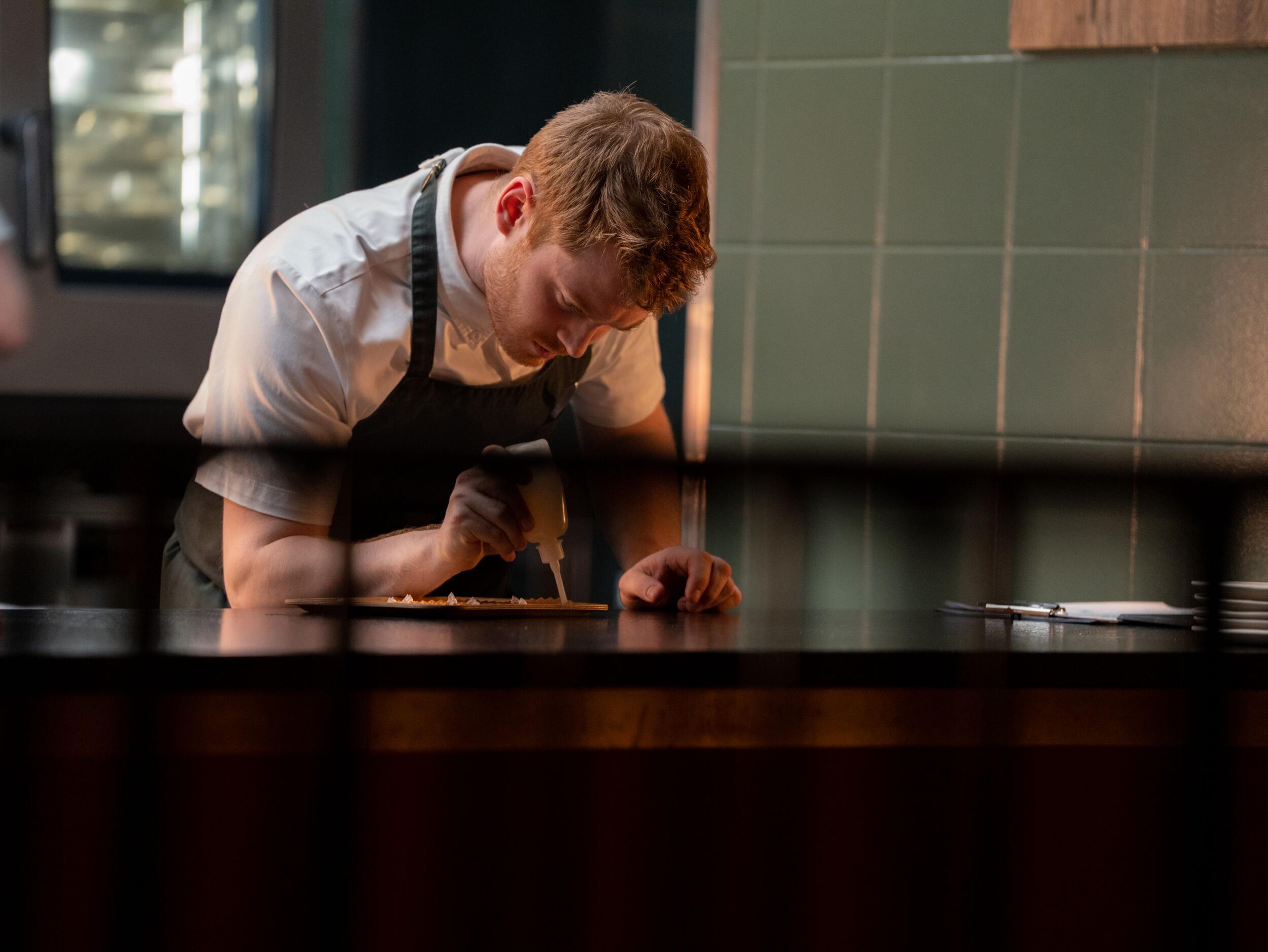 A chef intently plating food by drizzling sauce from a squeeze bottle.