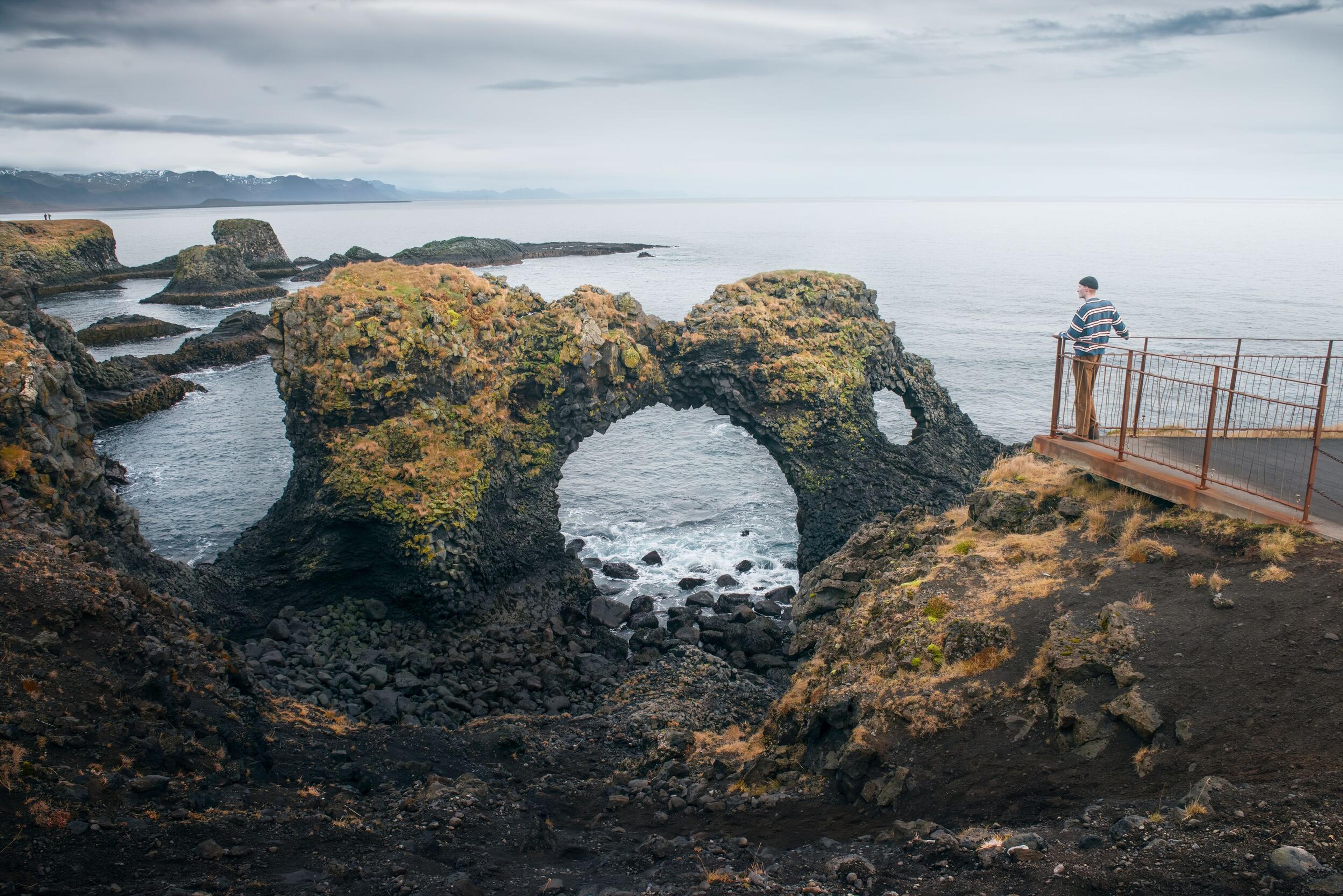 A person stands on a viewing platform overlooking a rugged coastline with distinctive rock arches and formations, under an overcast sky.