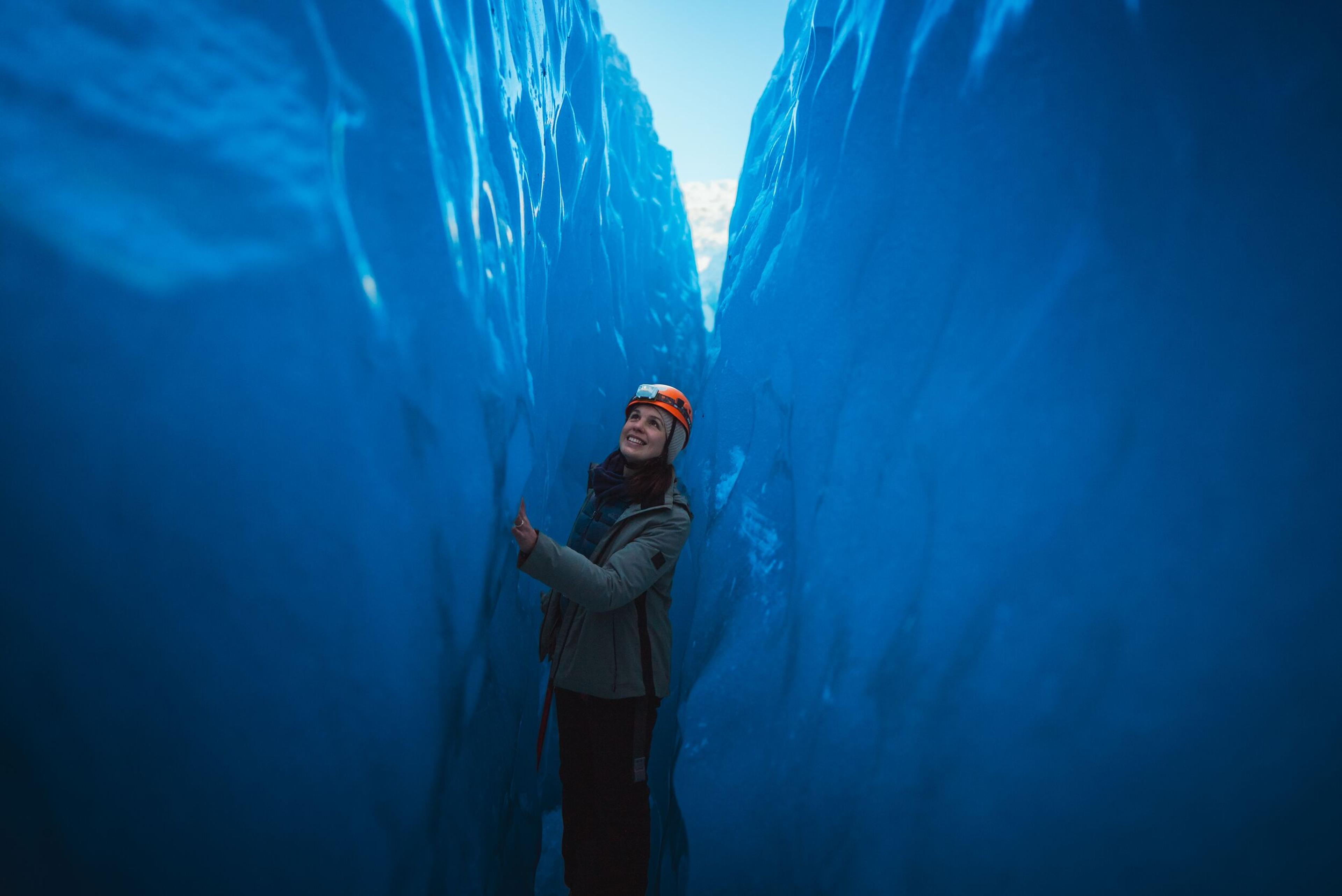 A woman in a helmet stands in a narrow blue ice cave, looking up.