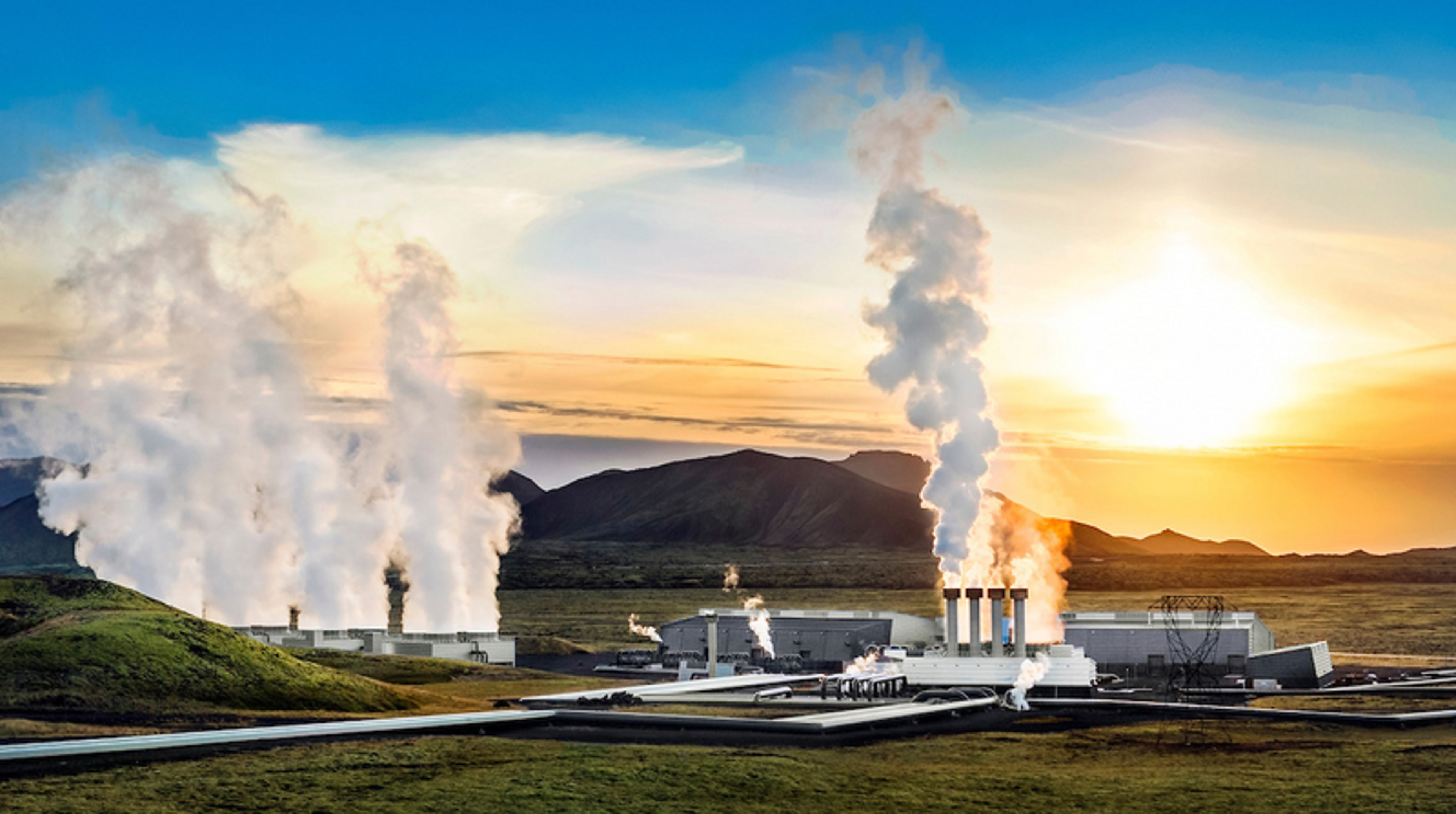 Sunset view of the Hellisheiði Power Plant in Iceland, featuring multiple steam columns rising against a vibrant sky, with the rugged landscape of volcanic hills in the background.
