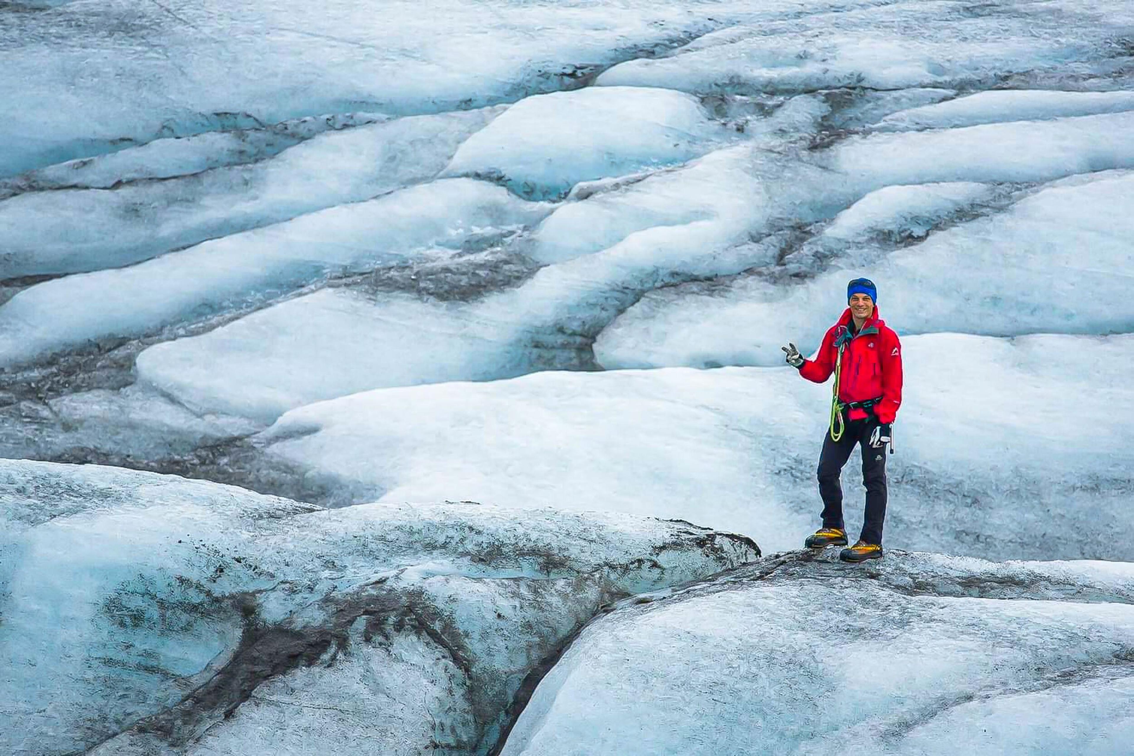 A lone hiker in red stands on the undulating, icy surface of a glacier, accentuating the vastness of the frozen landscape.