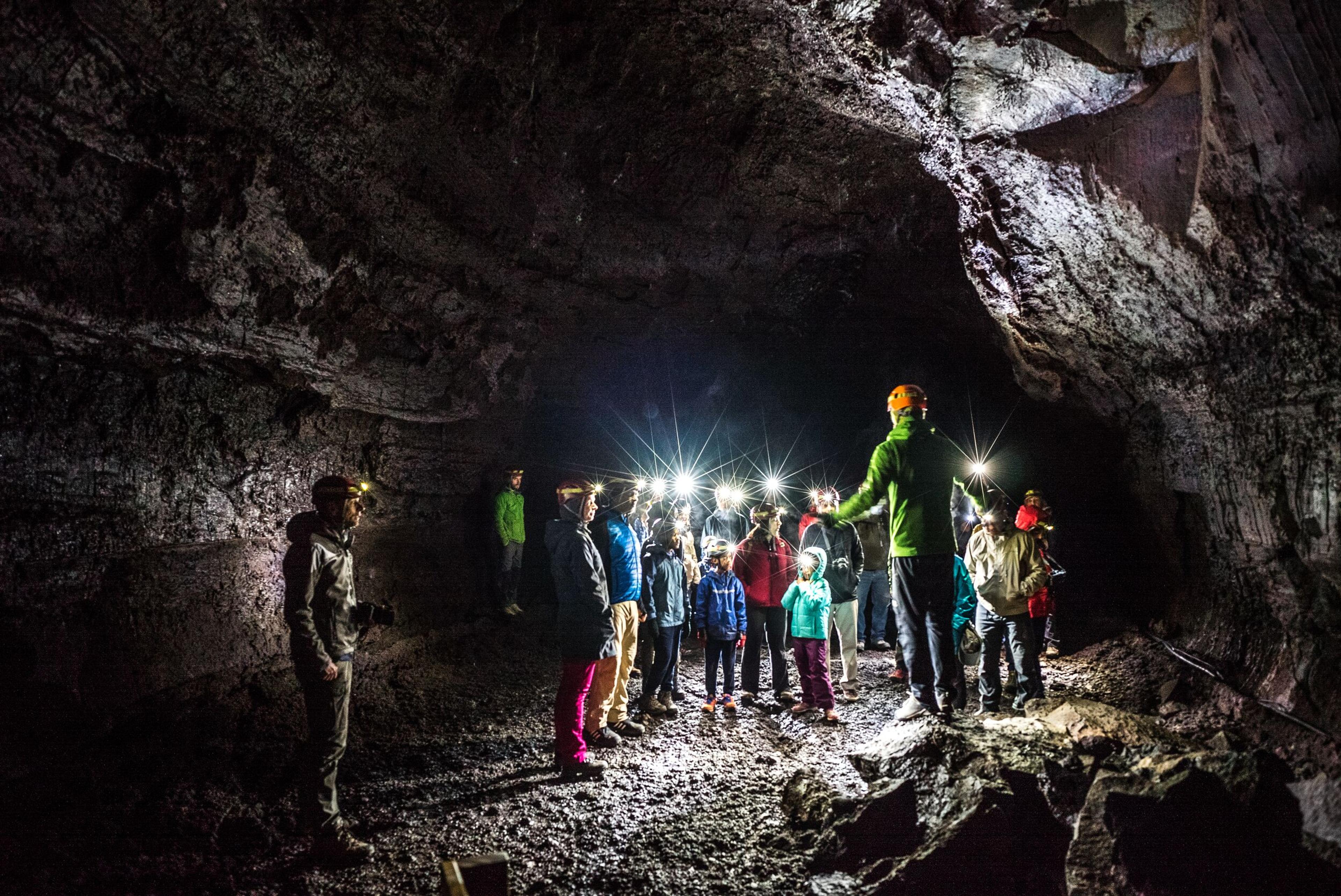 A group of tourists with headlamps exploring a large, dimly lit cave, guided by a tour leader wearing a helmet and bright jacket.