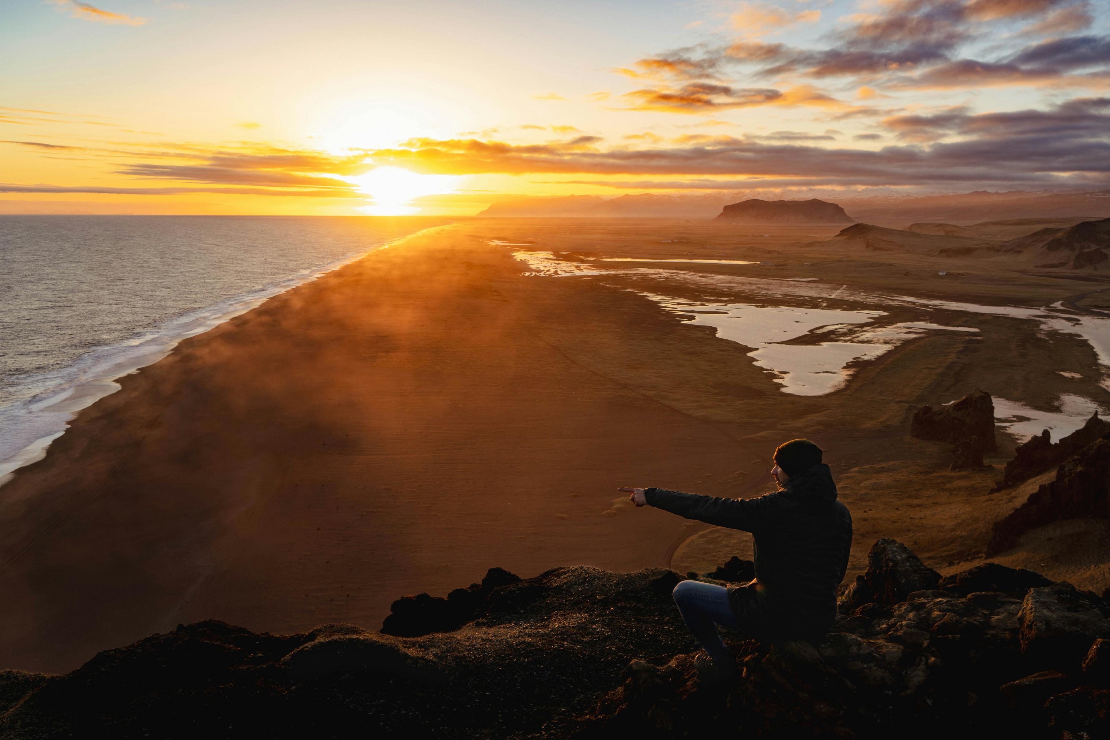 Person sitting on a cliff at Dyrhólaey, pointing towards the ocean at sunset, with a view of the coastline and mist rising from the beach.