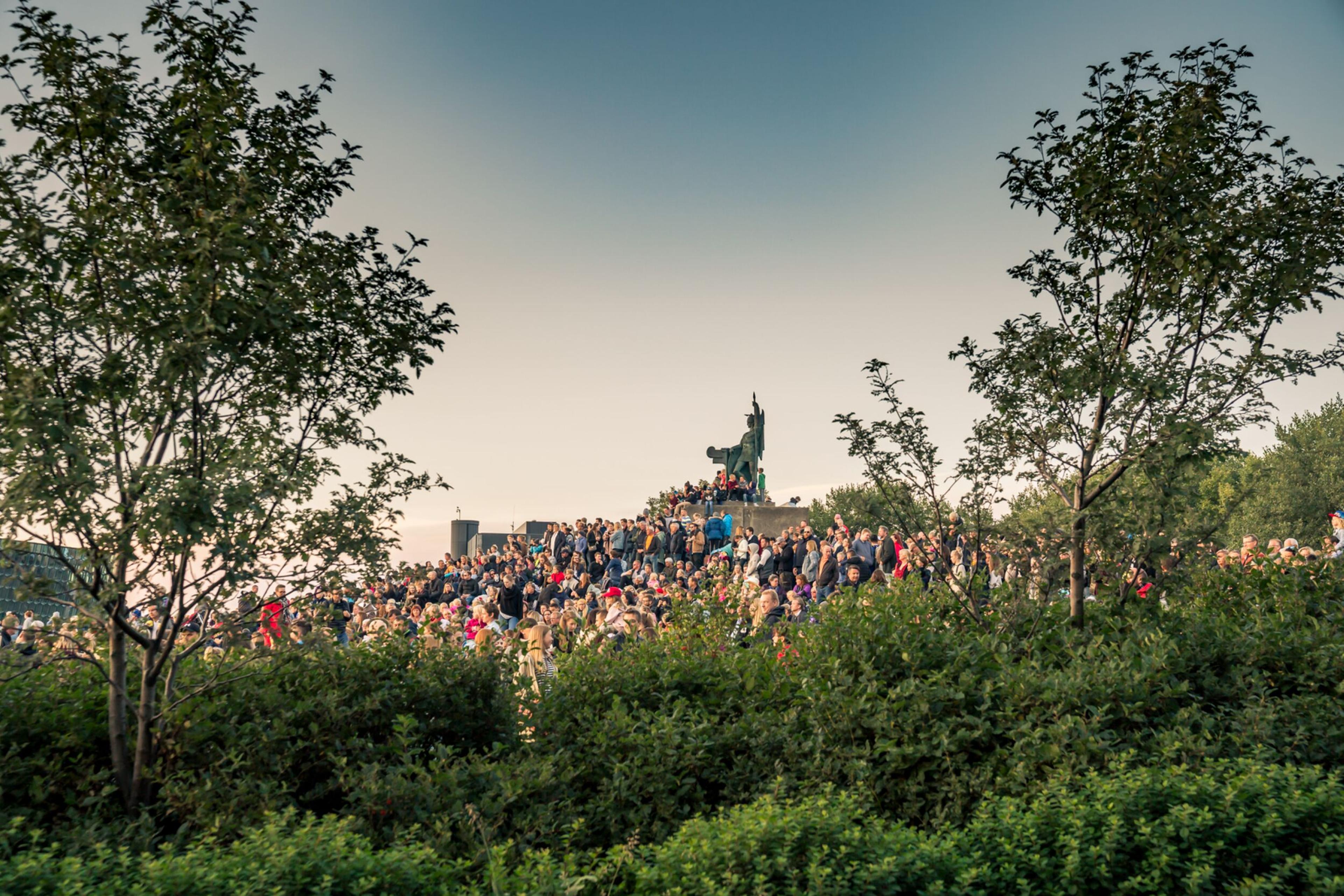 A large crowd gathers around a statue in a lush, green park during Reykjavík Culture Night, framed by trees under a clear evening sky.