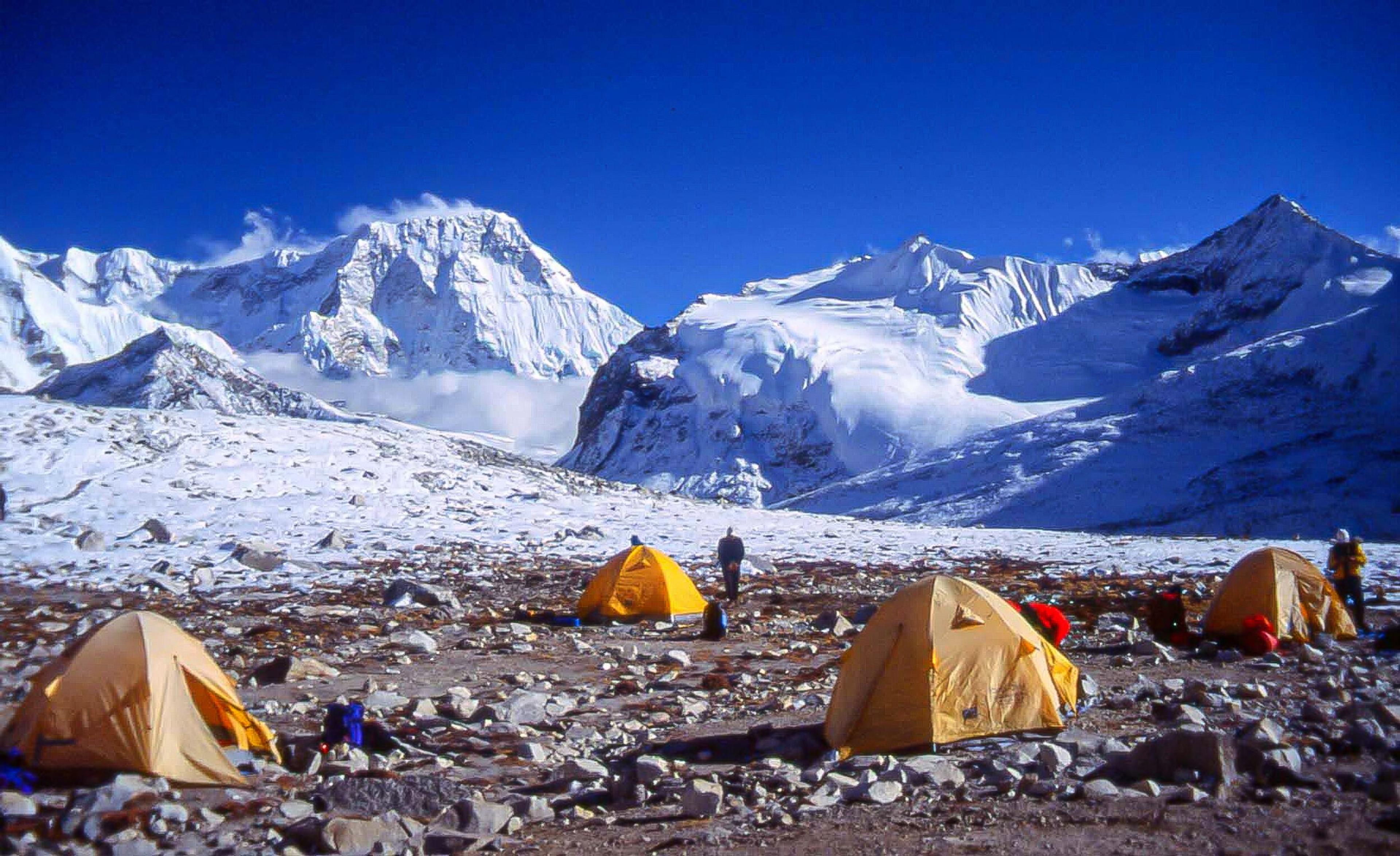 Orange tents dot a rugged base camp, dwarfed by the towering, snow-capped peaks stretching into the clear blue sky above.