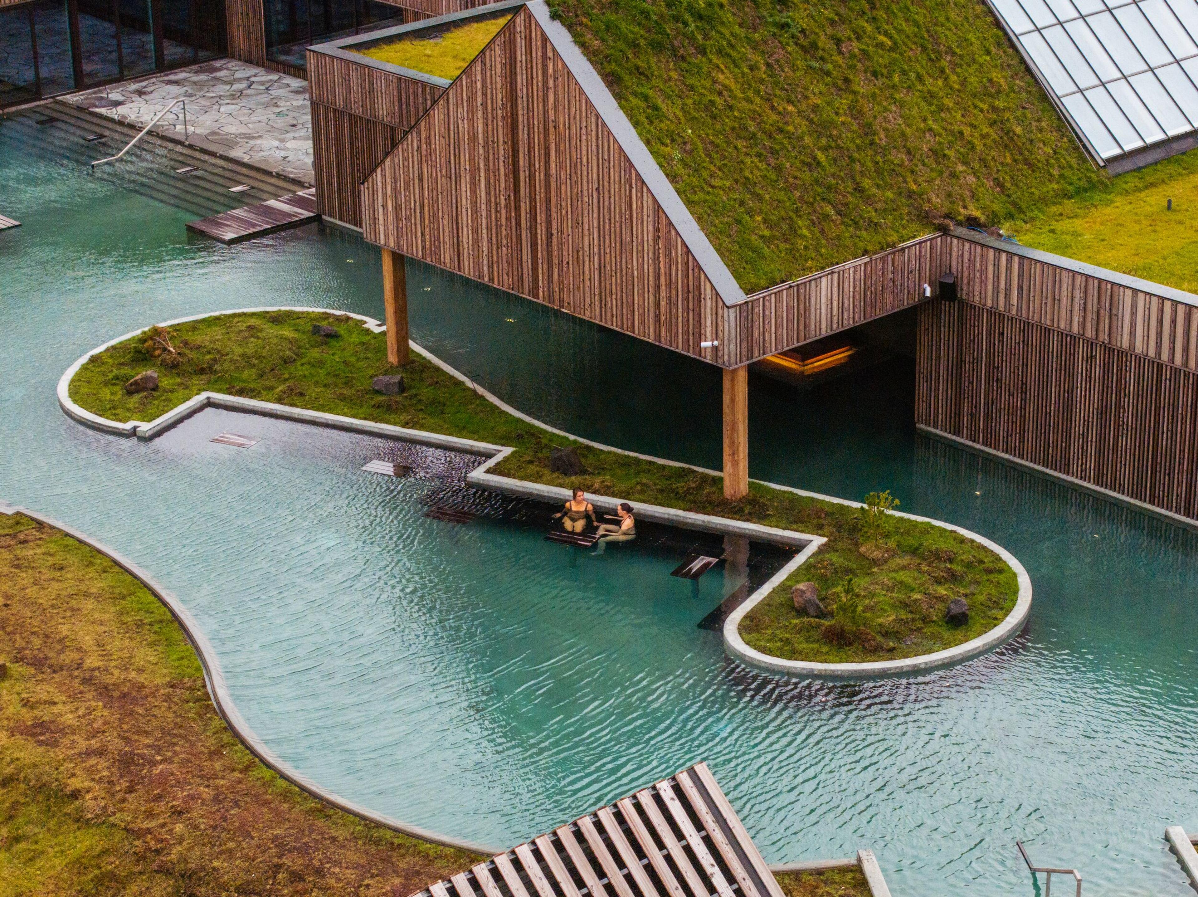 Modern thermal spa with green roofs, wooden buildings, and a winding blue pool containing two people.