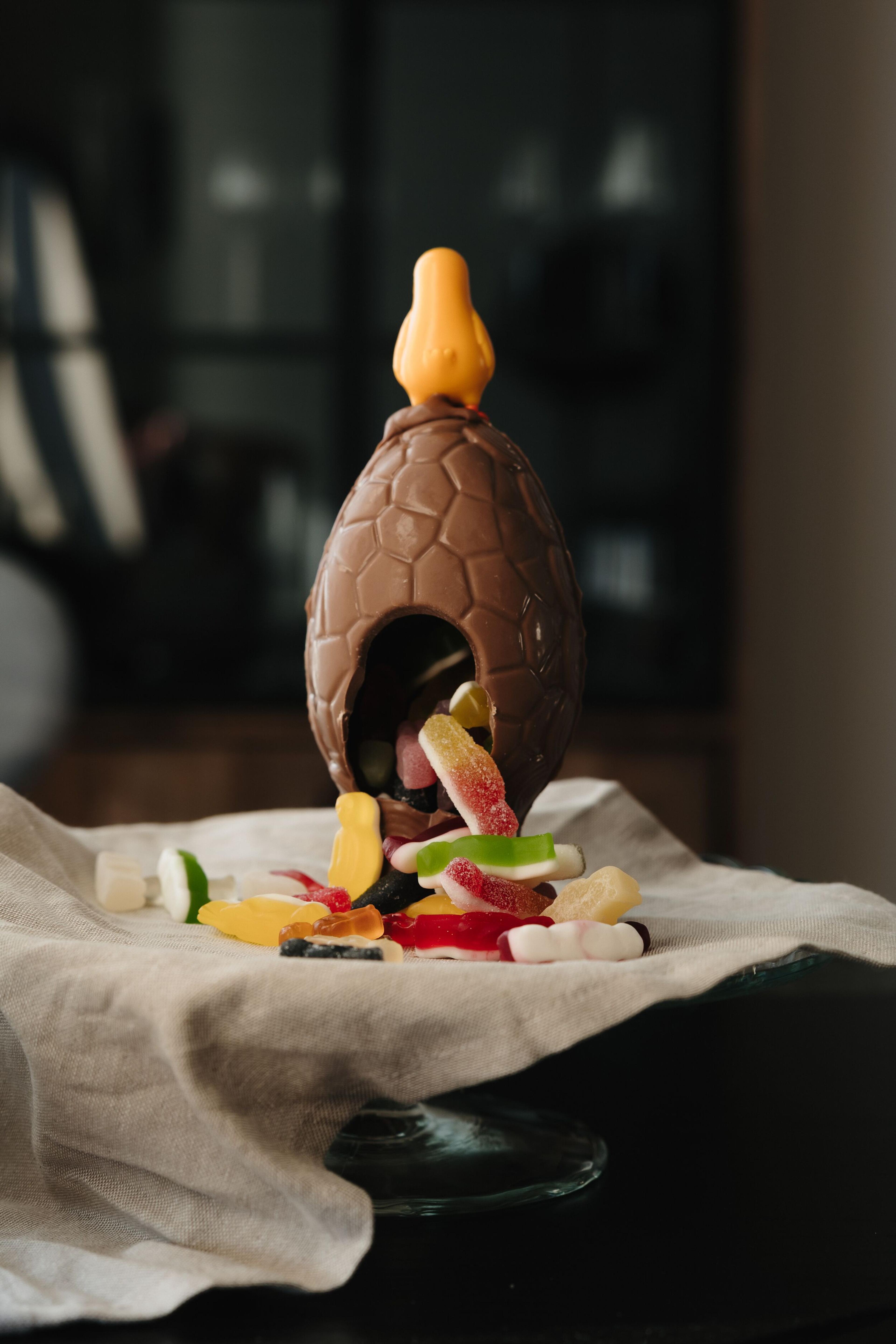 Textured chocolate Easter egg with an orange chick on top, cracked open and spilling colorful candies onto a linen cloth.