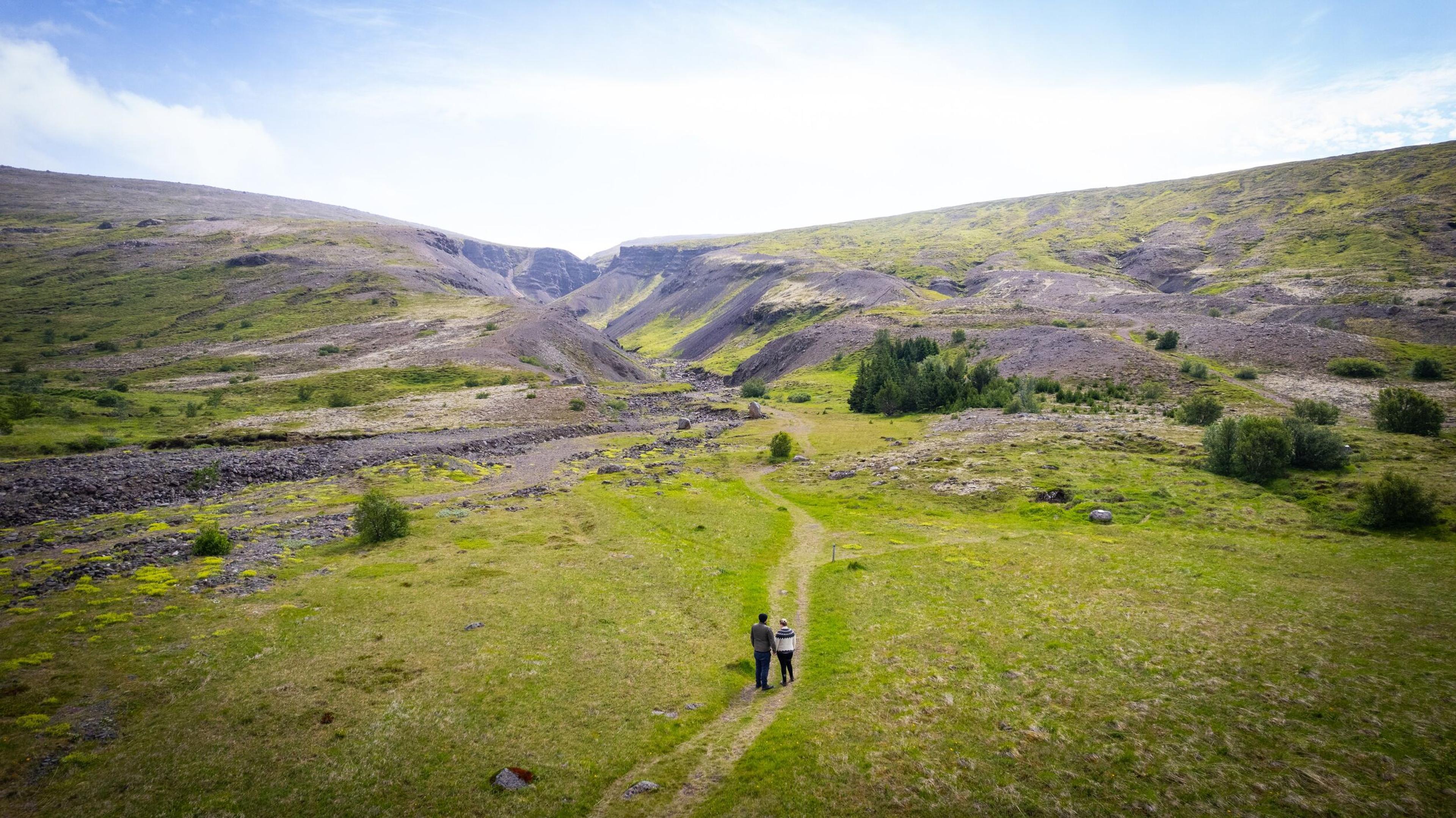 Two people walking on a narrow trail through a lush green valley surrounded by rocky hills and a distant gorge under a bright blue sky in Iceland.